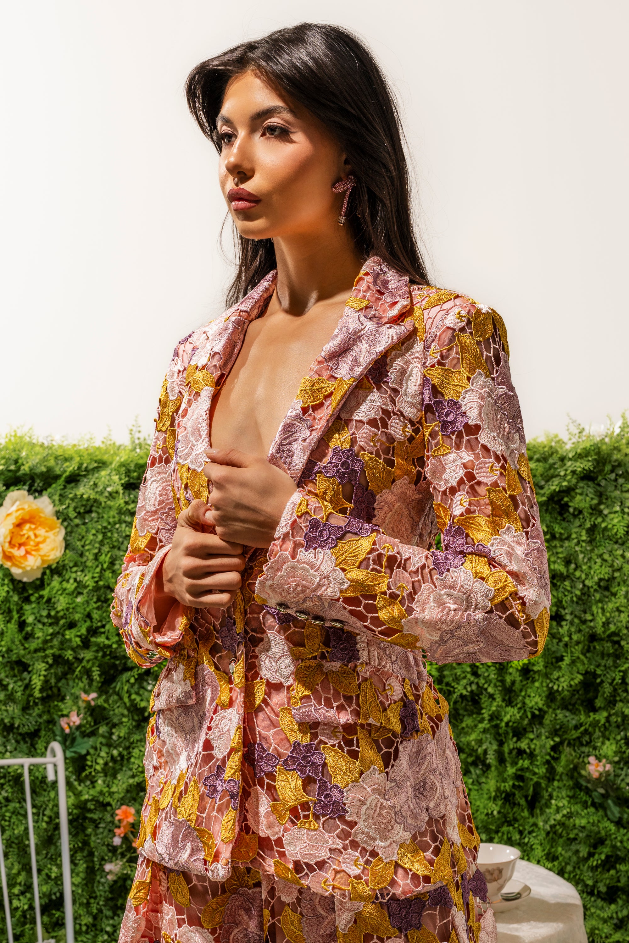 A woman with long dark hair wears the LILA LACE BLAZER, featuring colorful floral embroidery, as she stands before a flower-adorned green hedge and gazes thoughtfully to the side. 0