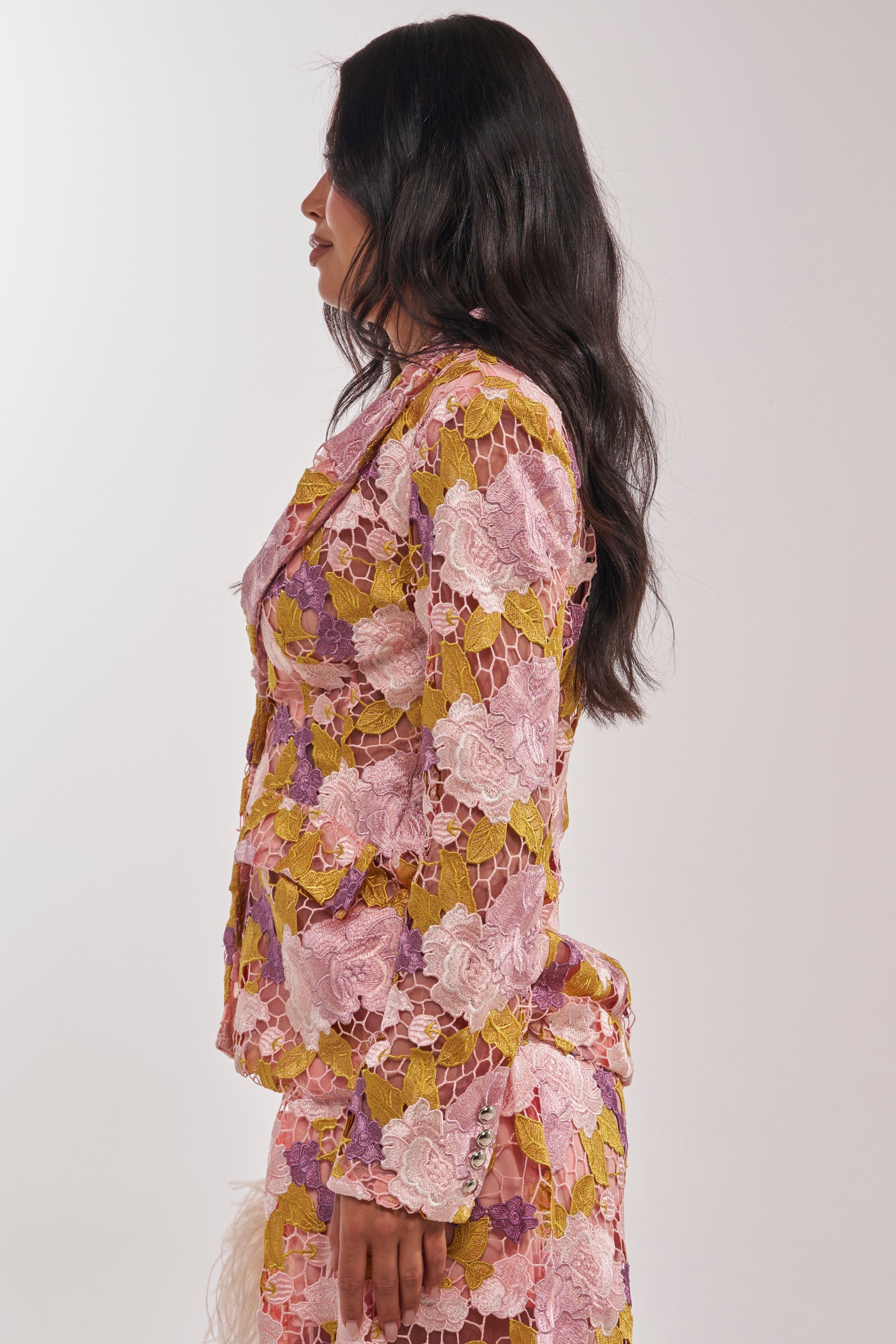A woman with long dark hair stands in profile against a plain light background, wearing the LILA LACE BLAZER—a colorful sheer blazer featuring pink, purple, and yellow floral lace patterns. 3