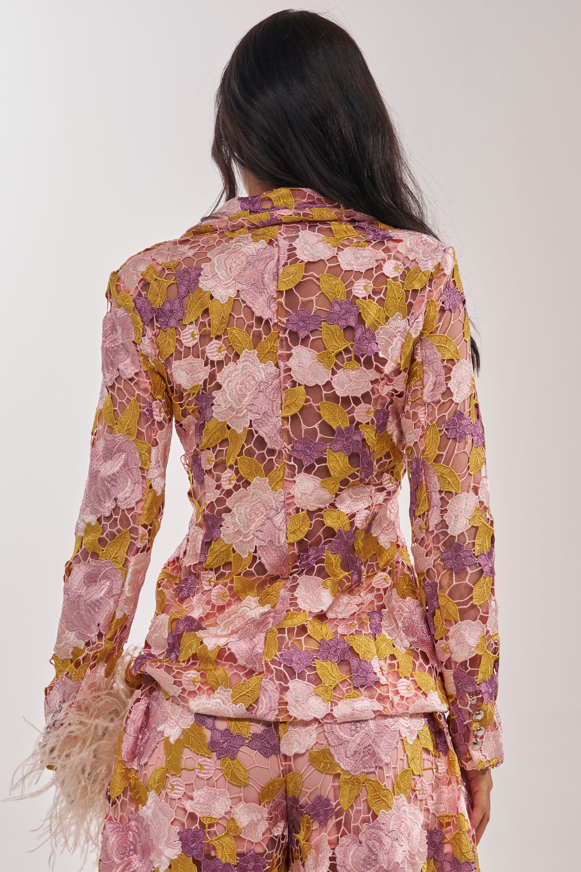 A woman with long dark hair faces away, showing the back of the LILA LACE BLAZER—a colorful sheer lace blazer and pants in pink, yellow, and purple with feathery sleeve details—against a plain background. 4