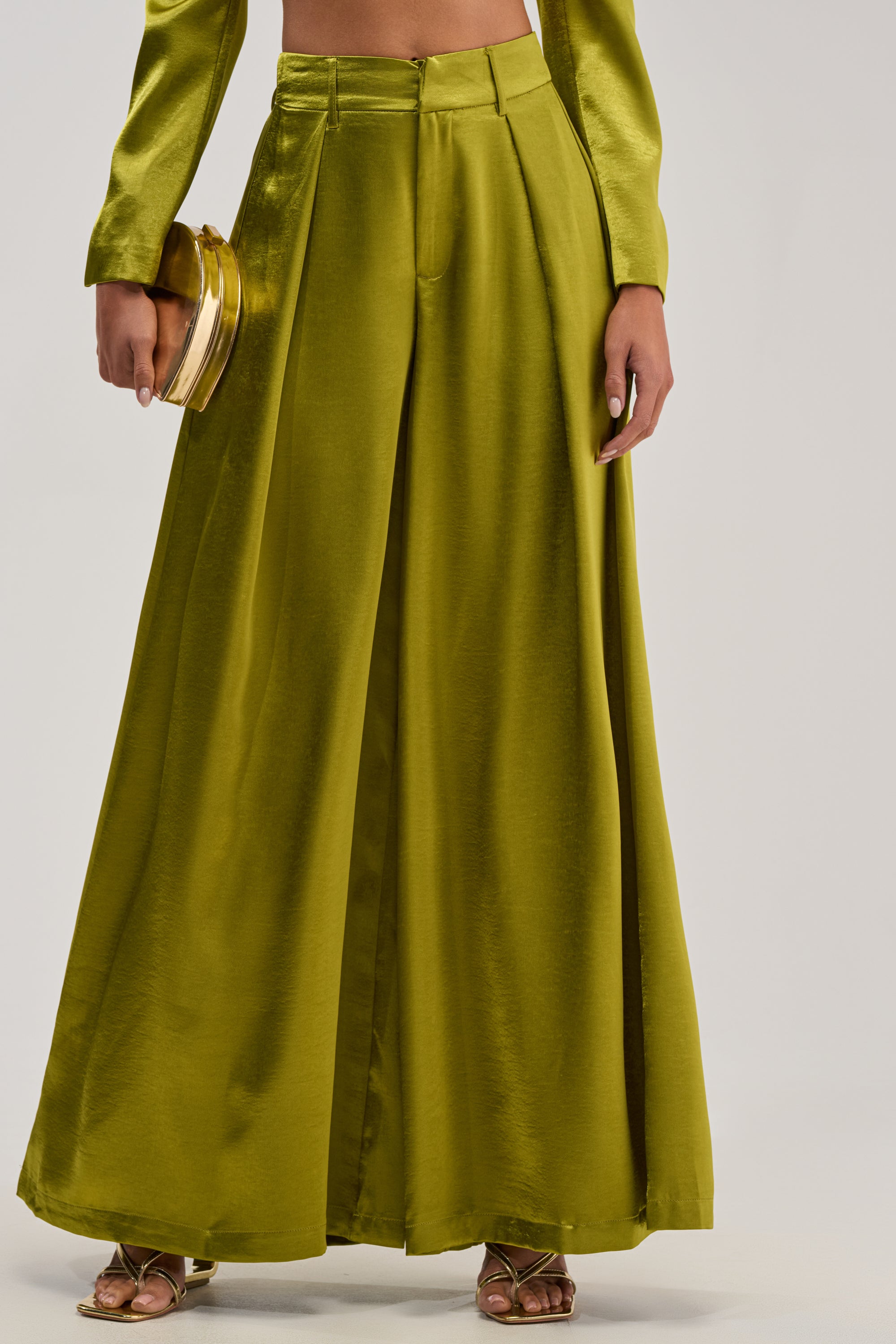 A person wearing the LUCKY ONE SATIN TROUSER in olive green with a matching long-sleeve crop top, gold heeled sandals, and a gold clutch—an ideal ensemble for formal evening events.
