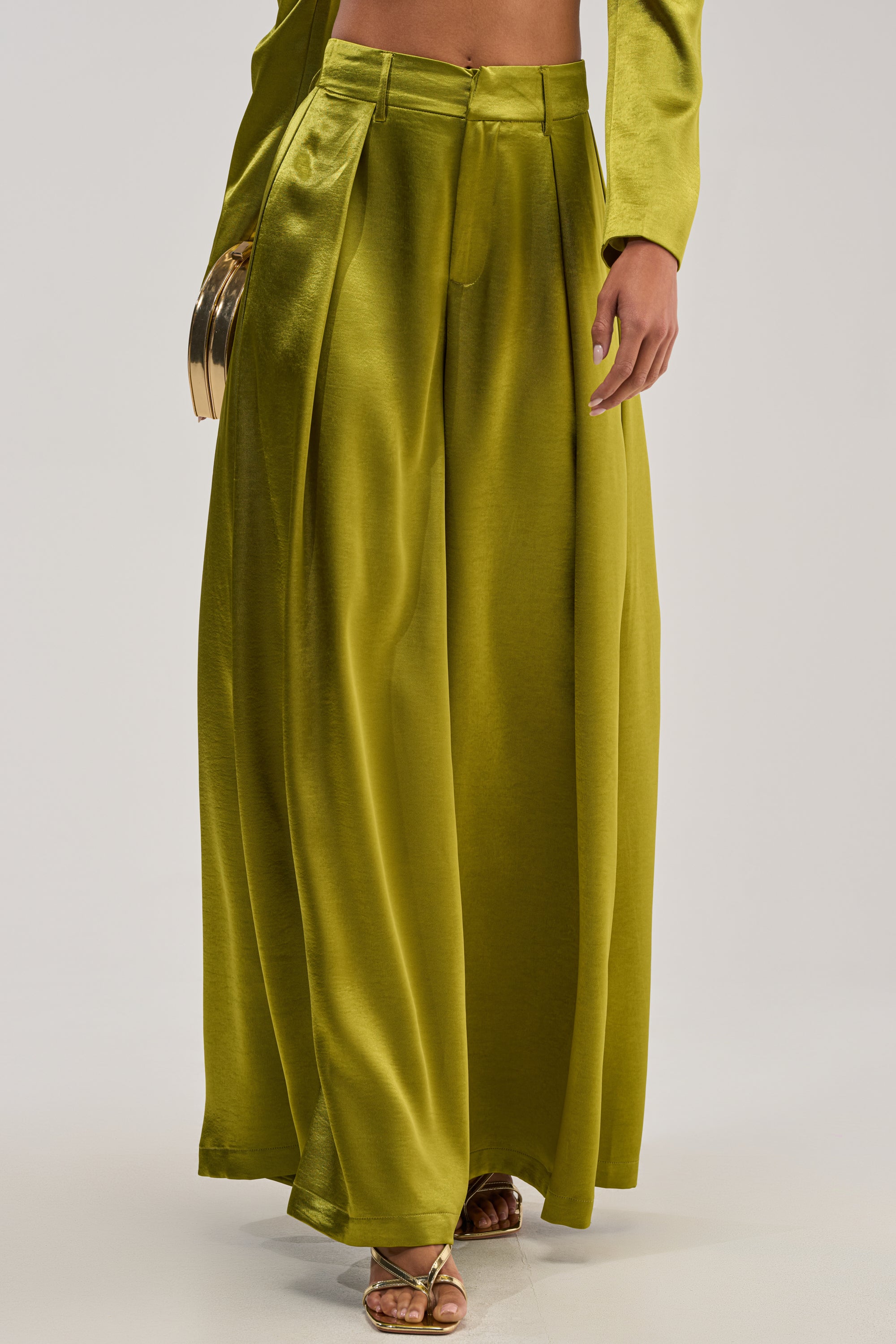 A person wears the LUCKY ONE SATIN TROUSER—shiny, high-waisted chartreuse green pants with pleats—in a wide-leg silhouette, styled with gold high-heeled sandals and a metallic clutch for an elegant formal evening look.