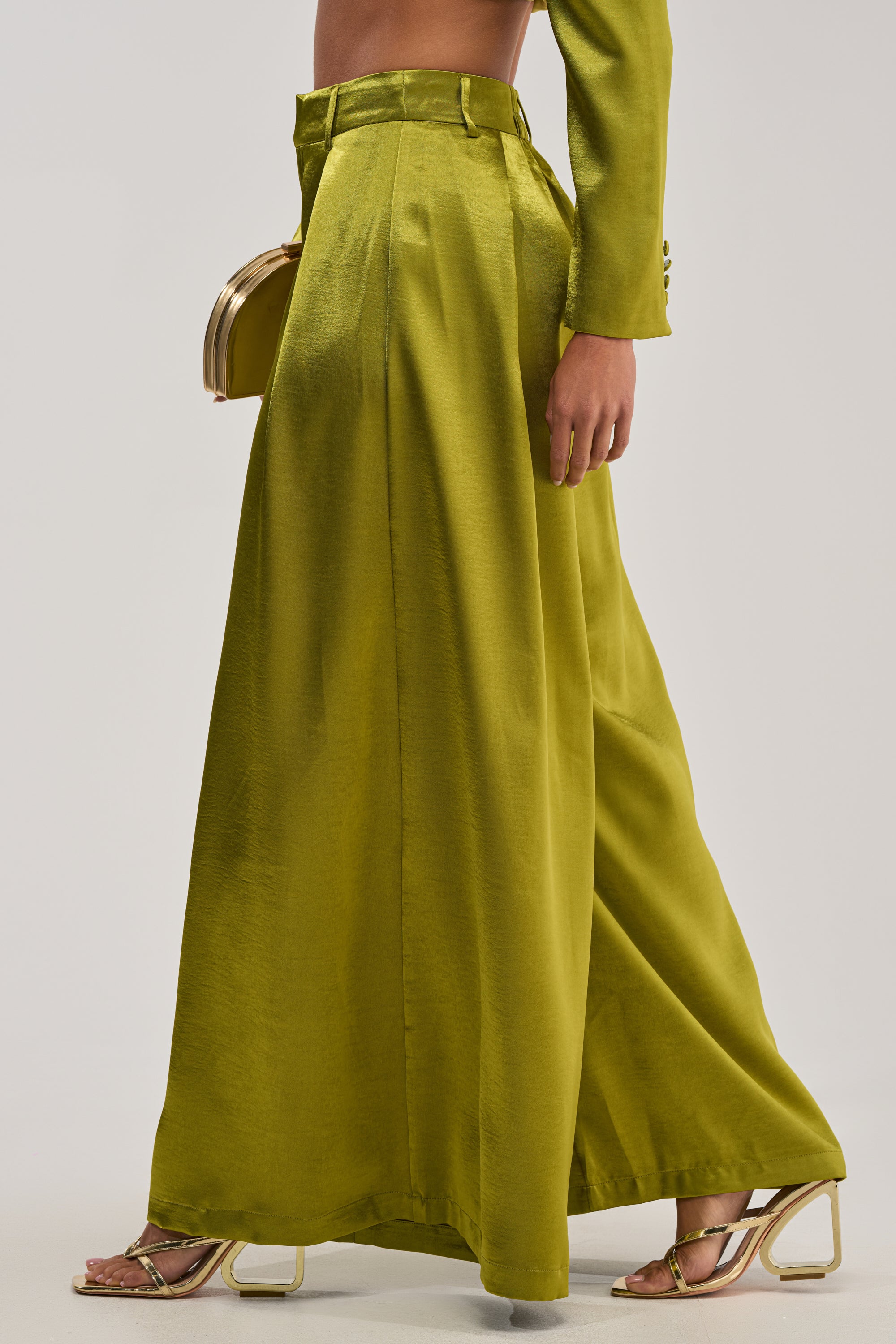 Dressed in the high-waisted LUCKY ONE SATIN TROUSER in green with a wide-leg silhouette, a matching blazer, metallic gold heels, and a gold clutch—an elegant look ideal for formal evening events.