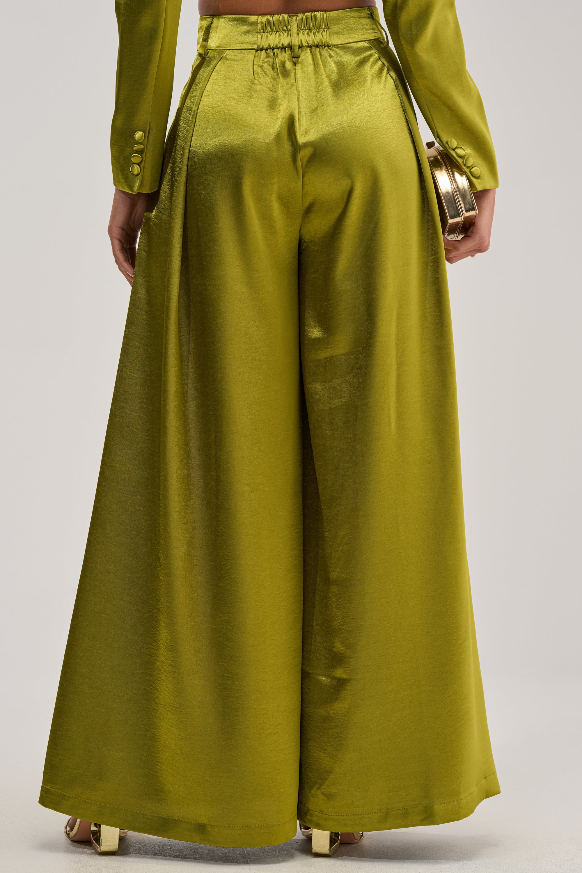 Wearing the LUCKY ONE SATIN TROUSER in shiny olive green with a matching jacket, a person holds a small gold clutch and is seen from behind against a neutral background.