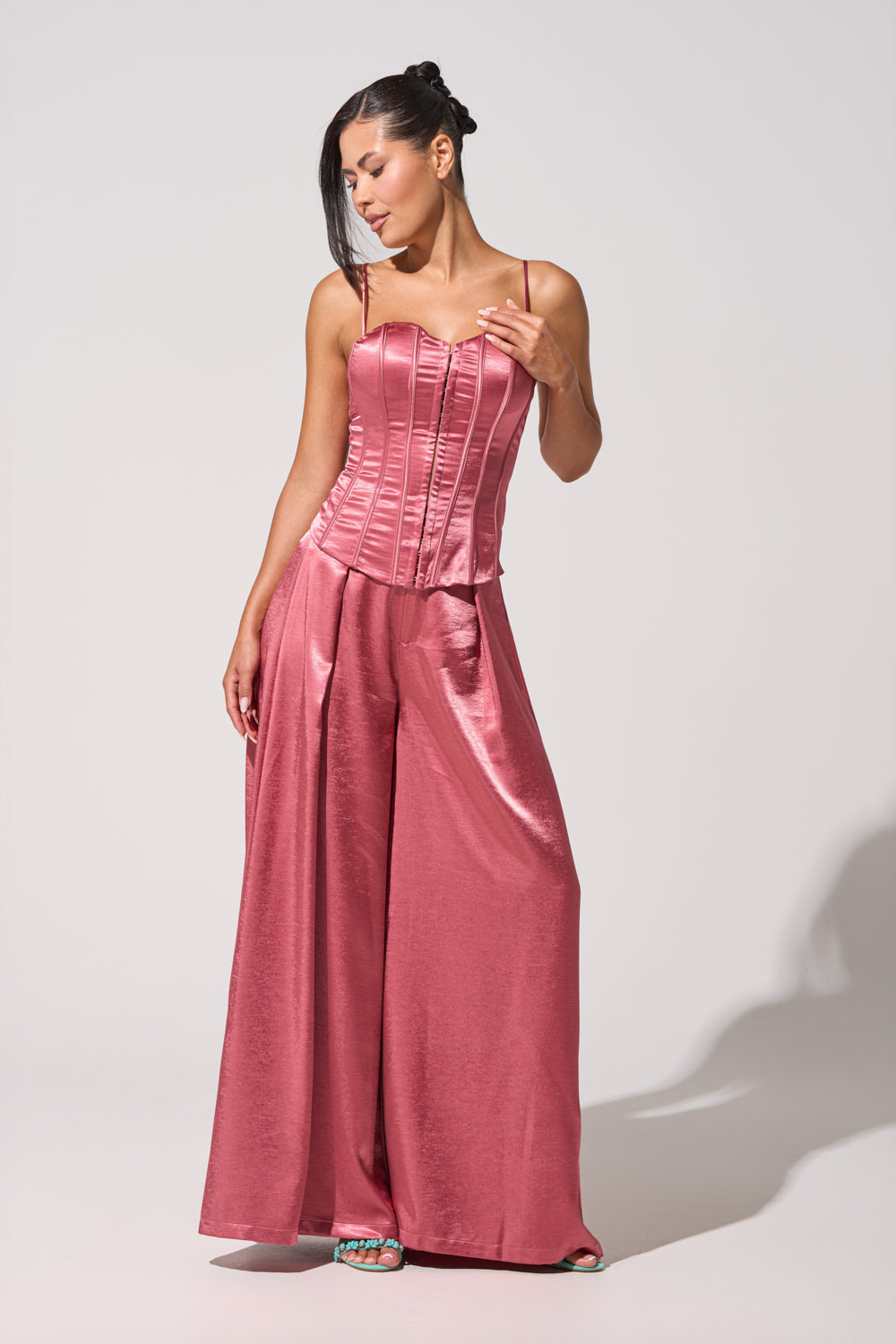 A woman stands in a light studio wearing the IN FULL BLOOM TROUSER IN MAUVE, a shiny pink satin high-rise pant paired with a fitted corset-style top, looking to the side and touching her hair. 1