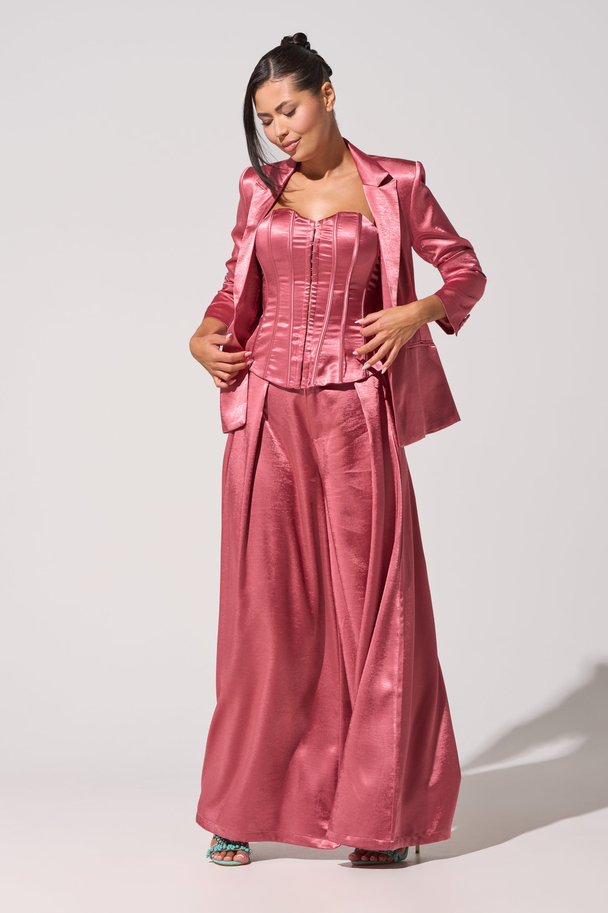 A woman stands against a plain light background, looking down as she touches her blazer while wearing the IN FULL BLOOM TROUSER IN MAUVE—shiny rose-pink high-rise satin wide-leg trousers styled with a matching blazer and corset top. 0