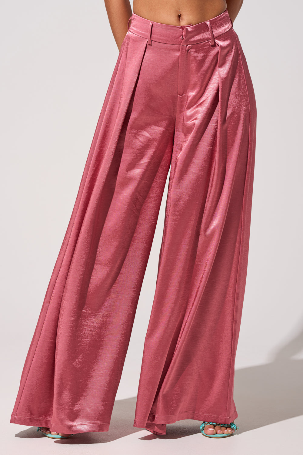 A person wears the IN FULL BLOOM TROUSER IN MAUVE—high-rise, shiny mauve satin wide-leg trousers with front pleats. Only their lower torso and legs are visible, paired with teal open-toe heels. 3