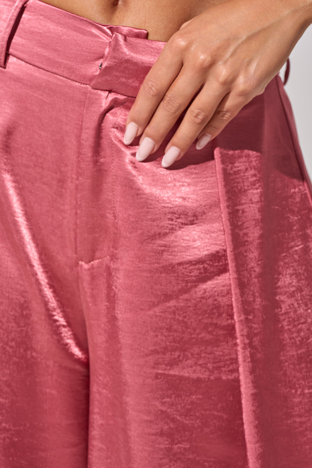 A close-up of a person wearing the IN FULL BLOOM TROUSER IN MAUVE, featuring shiny mauve satin fabric and a high-rise waist, with a hand resting on the hip and neatly manicured light pink nails. 8