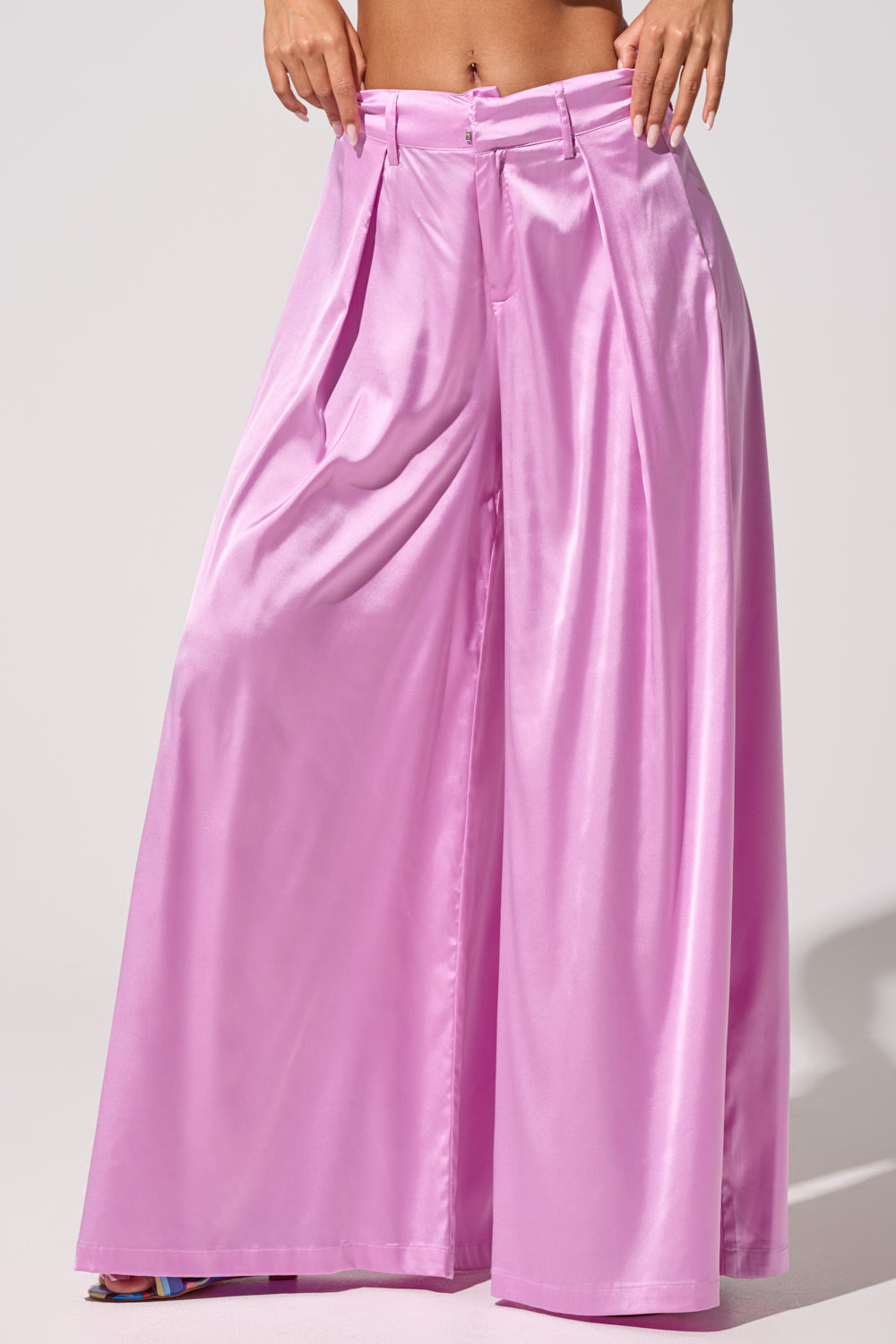 Wearing the IN FULL BLOOM LIGHT PURPLE TROUSER, a person stands against a plain light background with hands on their waist, showing off high-rise tailored evening pants in shiny satin and front pleats with a visible midriff. 2