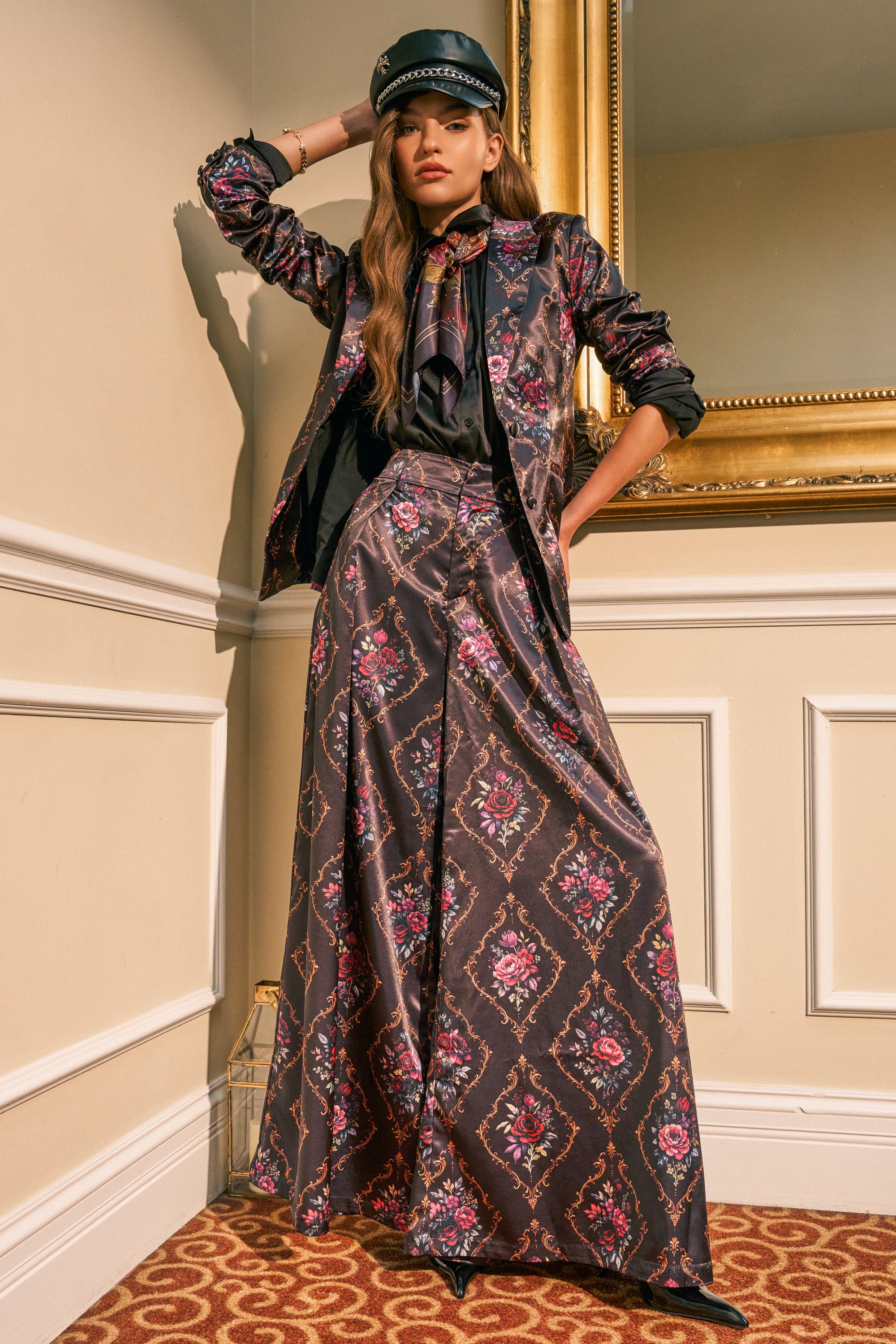 A woman poses indoors against cream-colored walls and a gold-framed mirror, wearing the VERTIGO SATIN TROUSER paired with a patterned blazer featuring an ornate floral print, a black shirt with a bow, and a black leather cap.