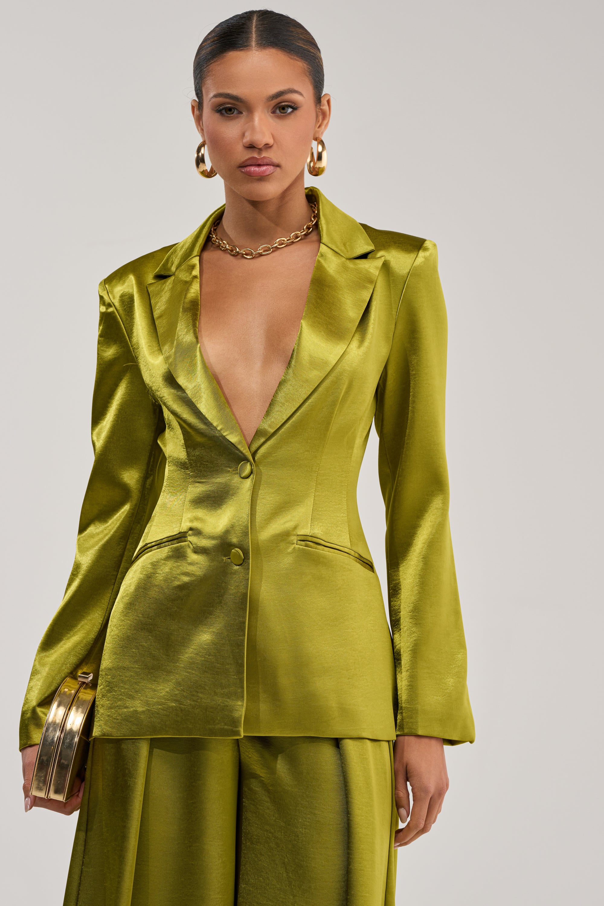 A woman models the LUCKY ONE SATIN BLAZER and matching green suit, featuring a deep V-neckline. She accessorizes with gold hoop earrings, a chunky necklace, and a round gold clutch—an ideal evening look for special occasions.