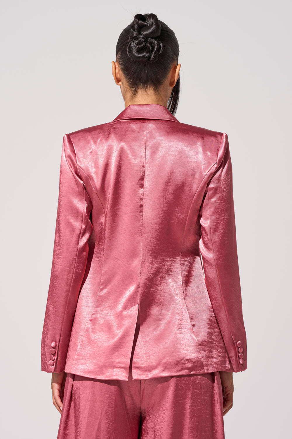 A person with dark hair in a neat bun models the IN FULL BLOOM BLAZER IN MAUVE, showcasing its shiny satin fabric and tailored silhouette from the back against a plain light background. 4