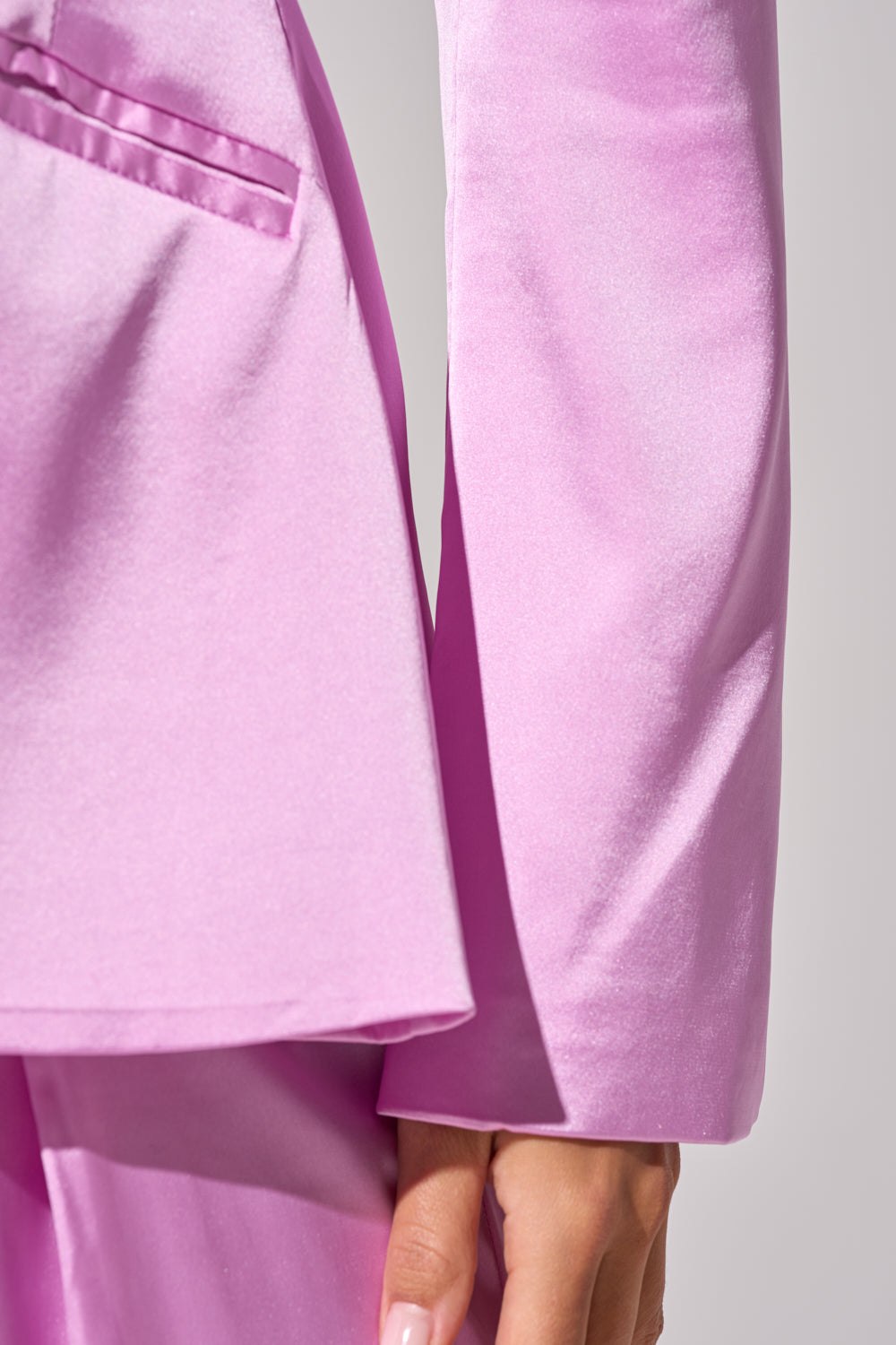 Close-up of a person wearing the IN FULL BLOOM LIGHT PURPLE BLAZER, highlighting the tailored satin sleeve, pocket, and pants against a neutral gray background with their hand visible at the bottom right. 6