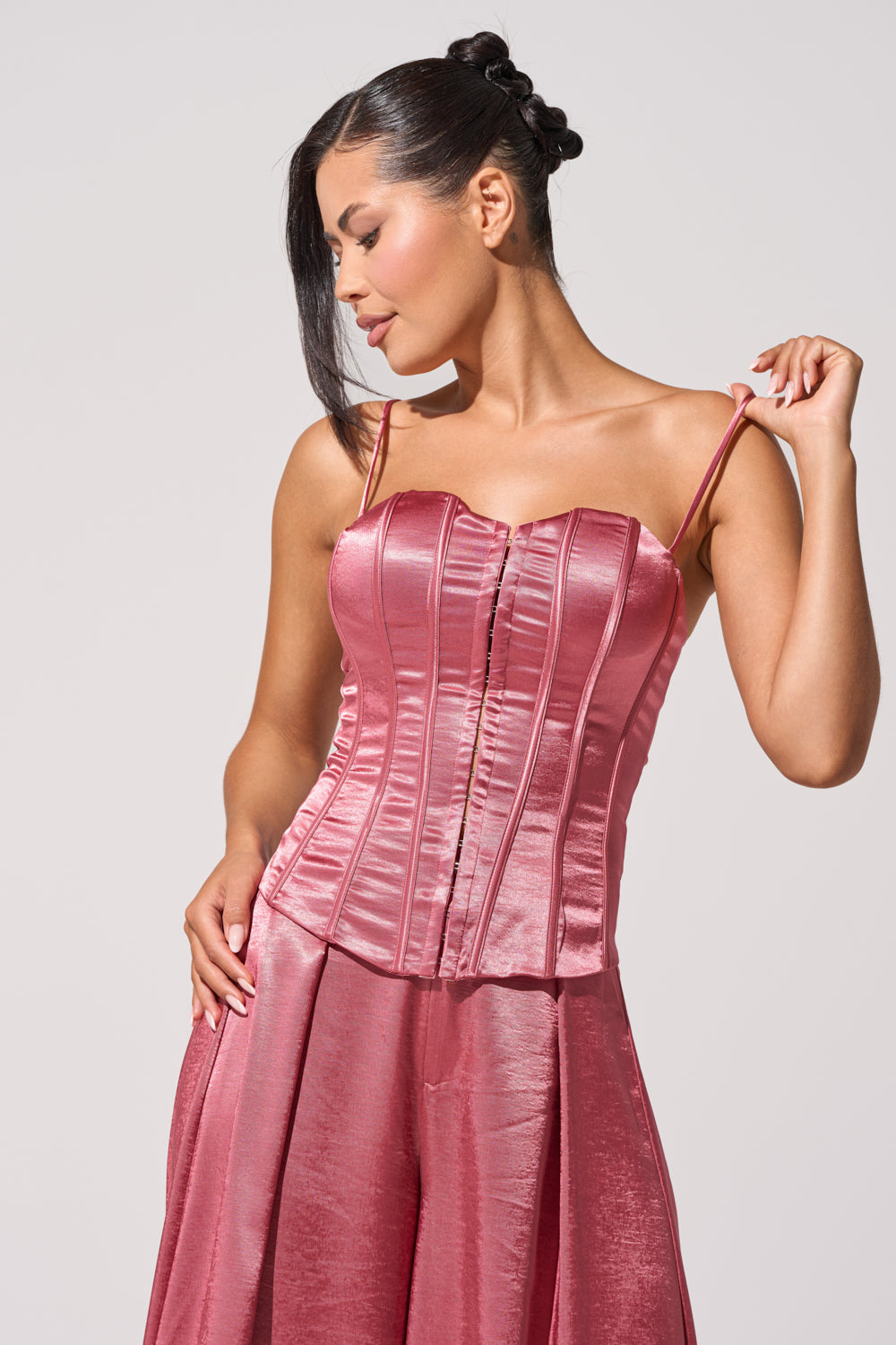 A woman poses confidently against a plain light background wearing the IN FULL BLOOM CORSET IN MAUVE, a shiny satin top with a sweetheart neckline and thin straps, paired with a matching skirt. 2