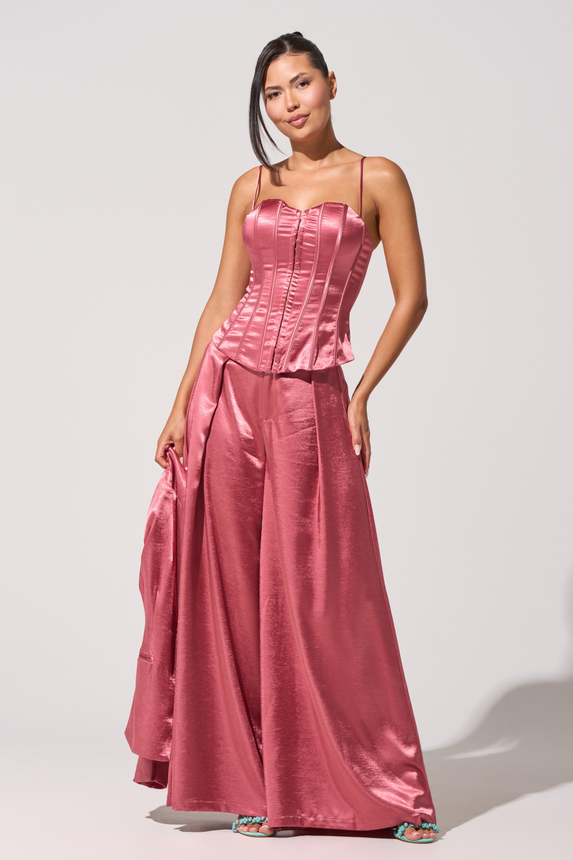A woman stands confidently in the IN FULL BLOOM CORSET IN MAUVE, a shiny satin top with structured boning. She pairs it with wide-leg pants and teal open-toe heels, holding matching fabric against a plain light gray background. 1