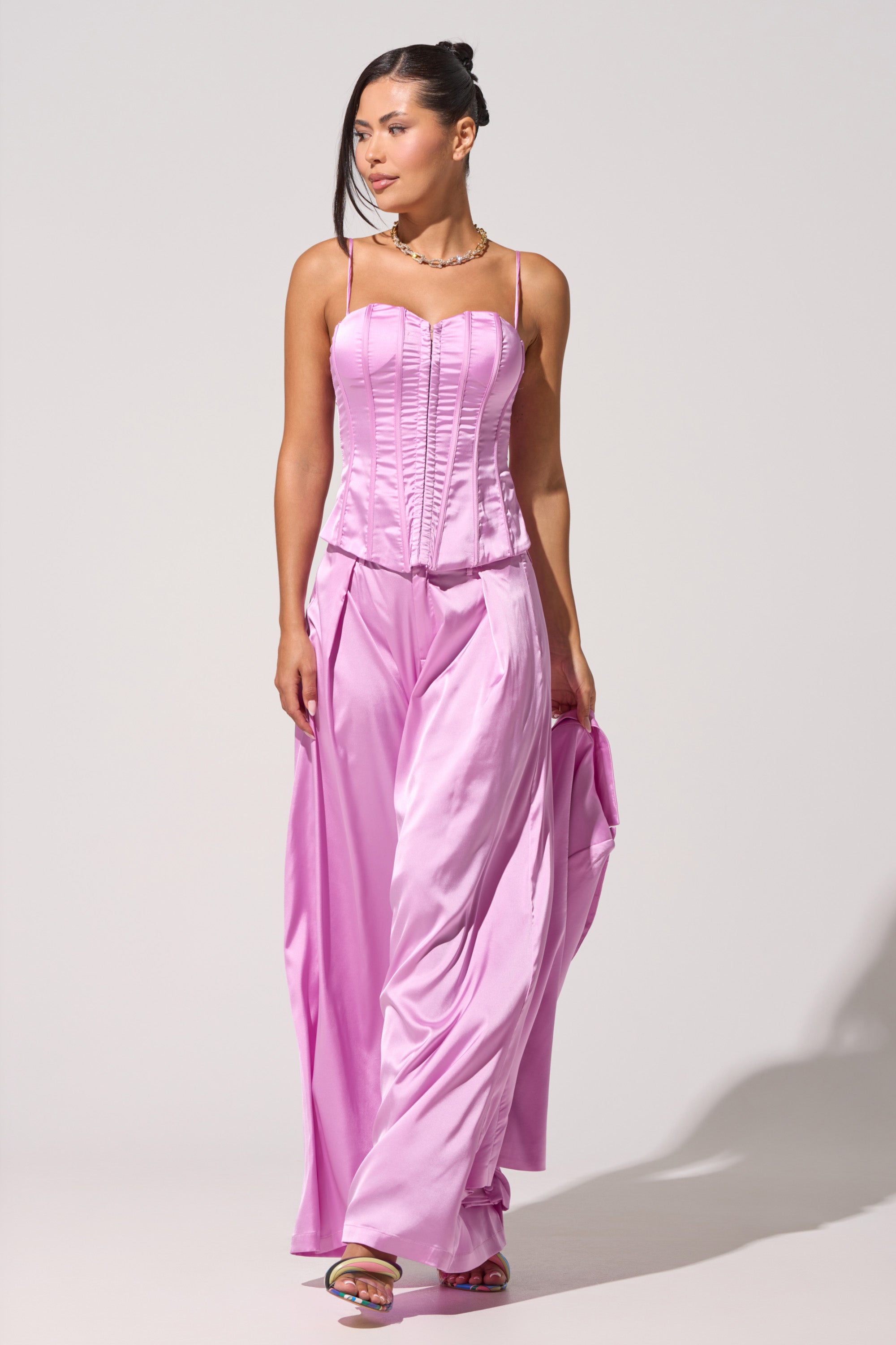 Wearing the IN FULL BLOOM LIGHT PURPLE CORSET with matching wide-leg pants, a woman stands against a plain background, holding her pants in one hand and gazing to the side—a stylish evening party look. 1