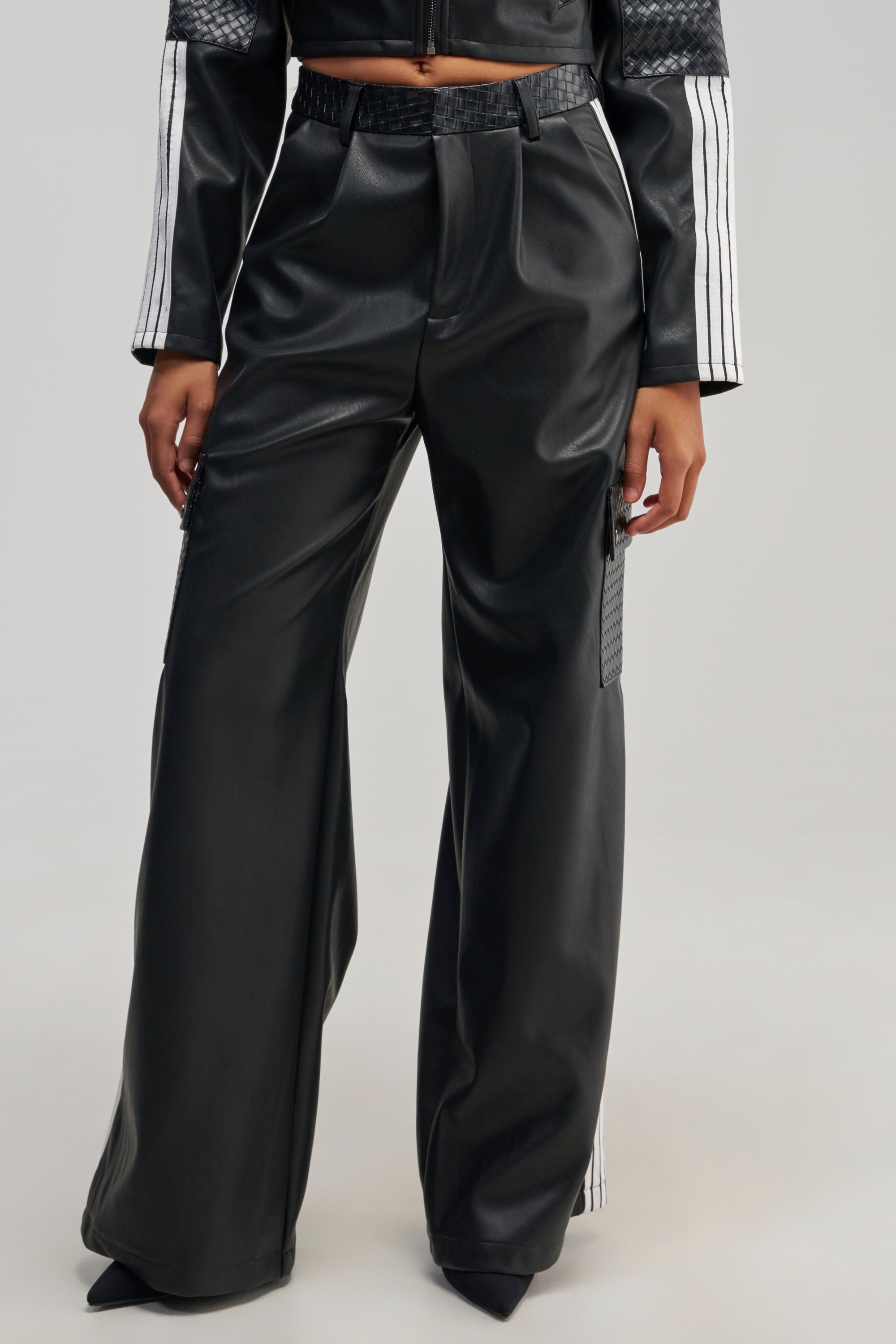 A person is wearing the DONT TEST ME FAUX LEATHER TROUSER in black, styled with a matching black top featuring bold white racing stripes on the sleeves, standing against a plain background.