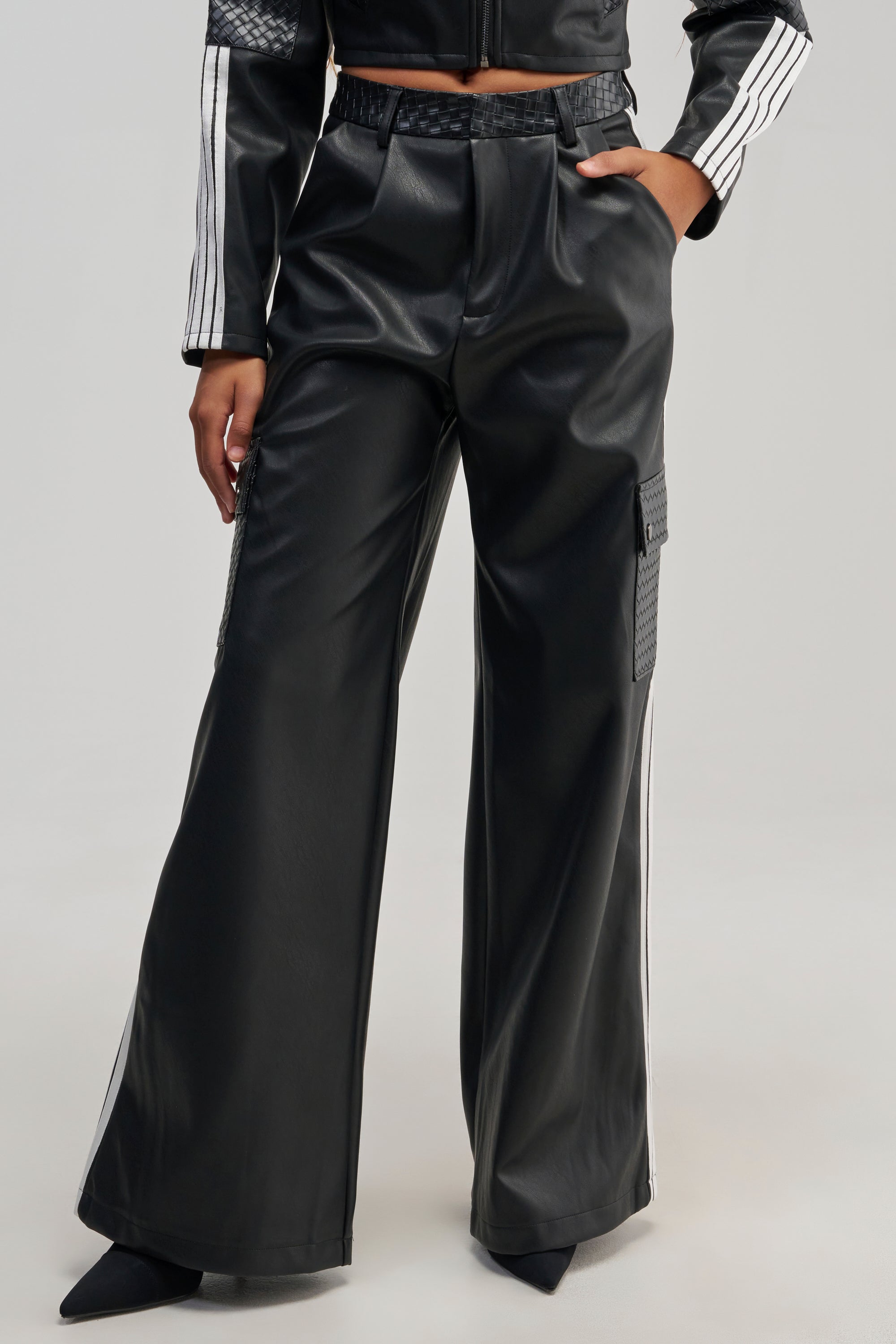 A person wearing the DONT TEST ME FAUX LEATHER TROUSER—black wide-leg faux leather cargo pants featuring bold white racing stripes along the sides and textured pockets—paired with a matching cropped jacket. Only the body from mid-torso down is visible.