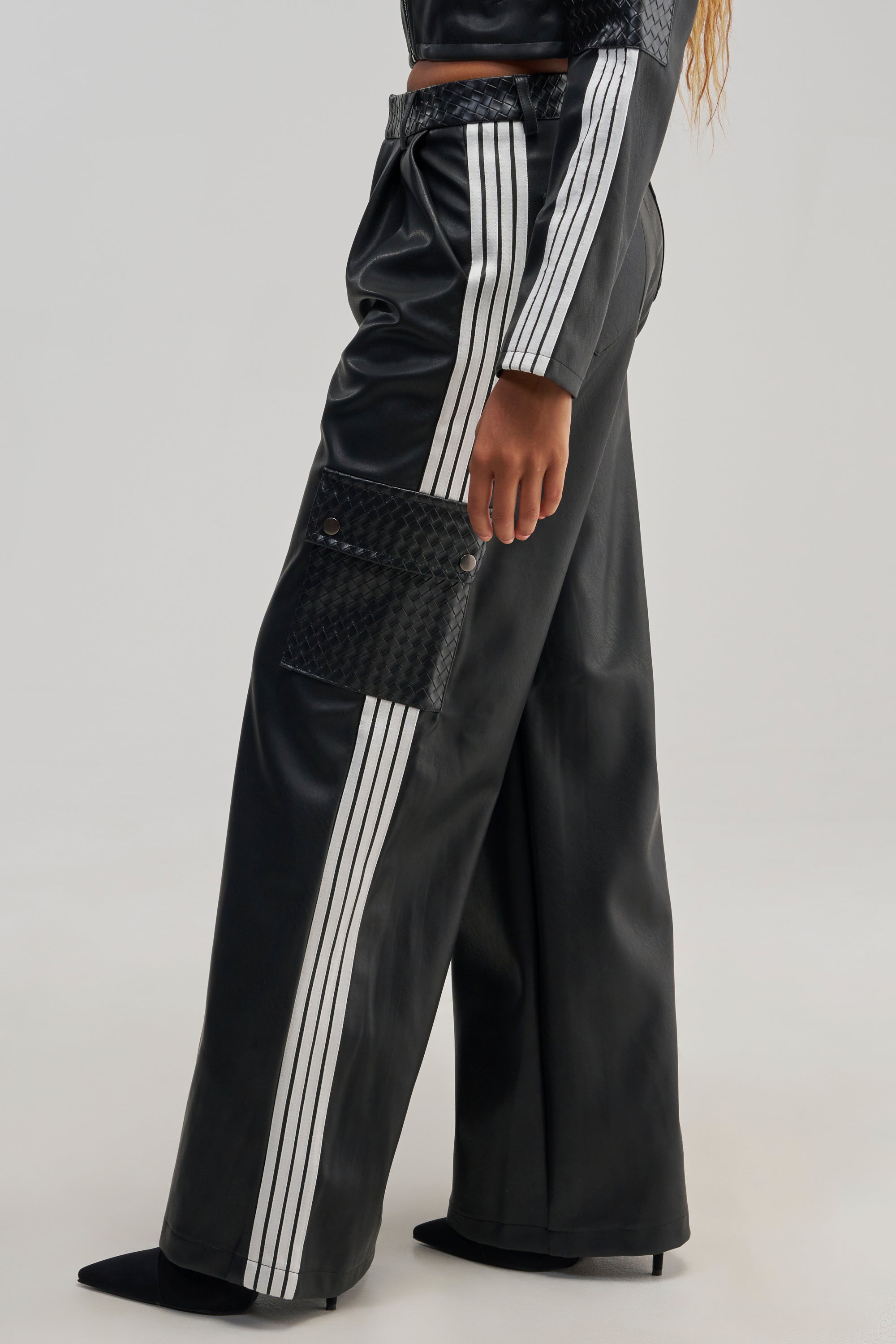 A person wearing the DONT TEST ME FAUX LEATHER TROUSER, featuring black faux leather with white racing stripes down the sides and a textured side pocket, paired with matching black heeled shoes and a cropped top.