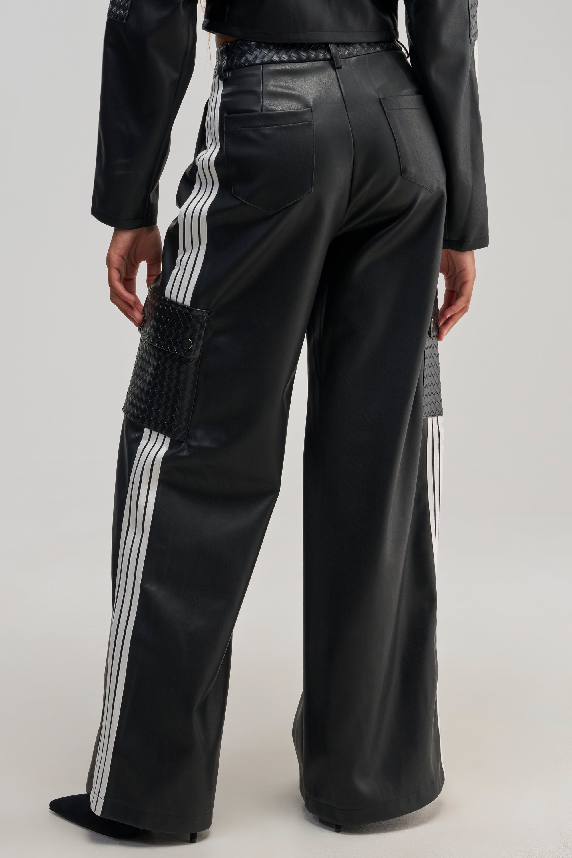 A person is seen from the back wearing the DONT TEST ME FAUX LEATHER TROUSER—black faux leather pants featuring racing-inspired white stripes down the sides and textured black thigh patches. The wide-leg fit adds a sporty edge, styled here with a coordinating black leather jacket.