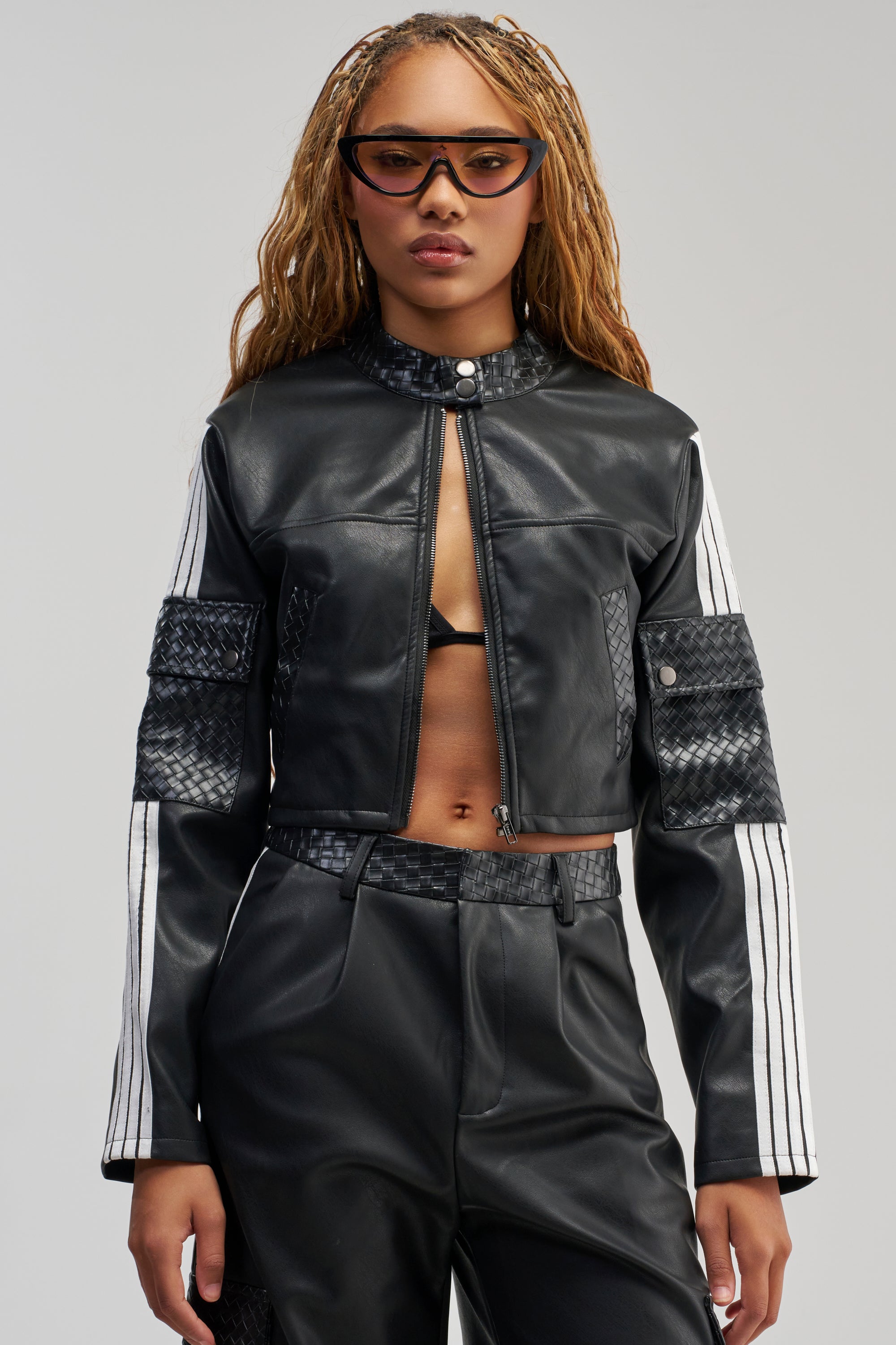 A person with long, braided blond hair wears cat-eye sunglasses and the DONT TEST ME FAUX LEATHER BOMBER—a black faux leather jacket with white stripes on the sleeves—paired with matching leather pants, standing against a plain light background.