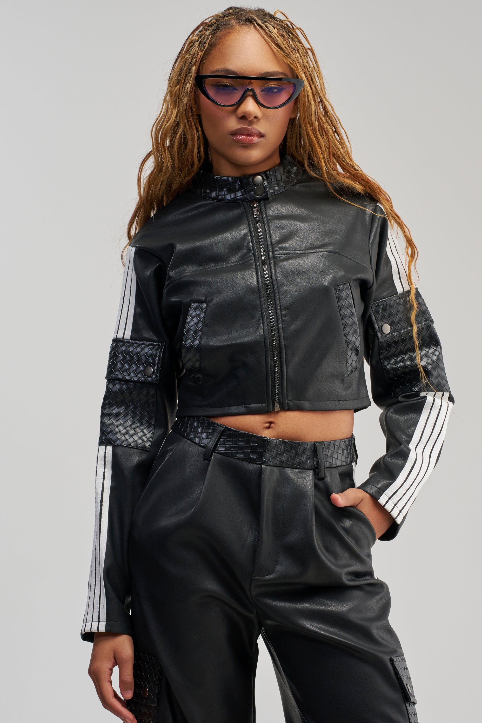 A woman with long braids poses confidently against a plain background, wearing the DONT TEST ME FAUX LEATHER BOMBER—a black faux leather jacket with white stripes on the sleeves—paired with matching high-waisted pants and purple sunglasses.