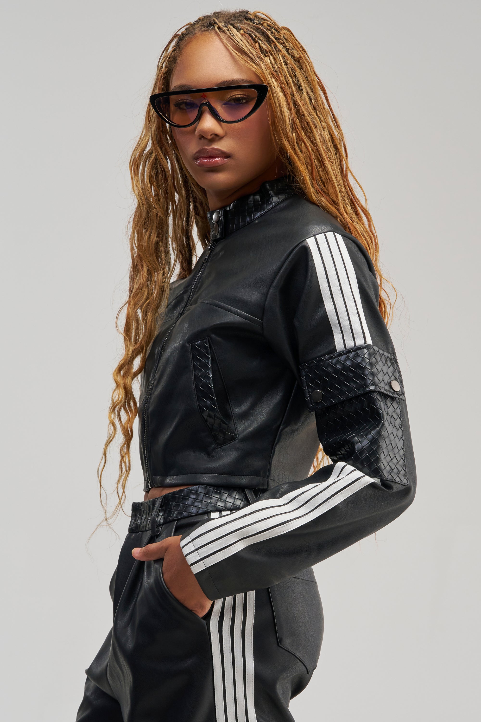 A person with long braided hair wears black sunglasses and the DONT TEST ME FAUX LEATHER BOMBER over a black outfit featuring white stripes on the sleeves and pants. They stand confidently with one hand in their pocket against a plain background.