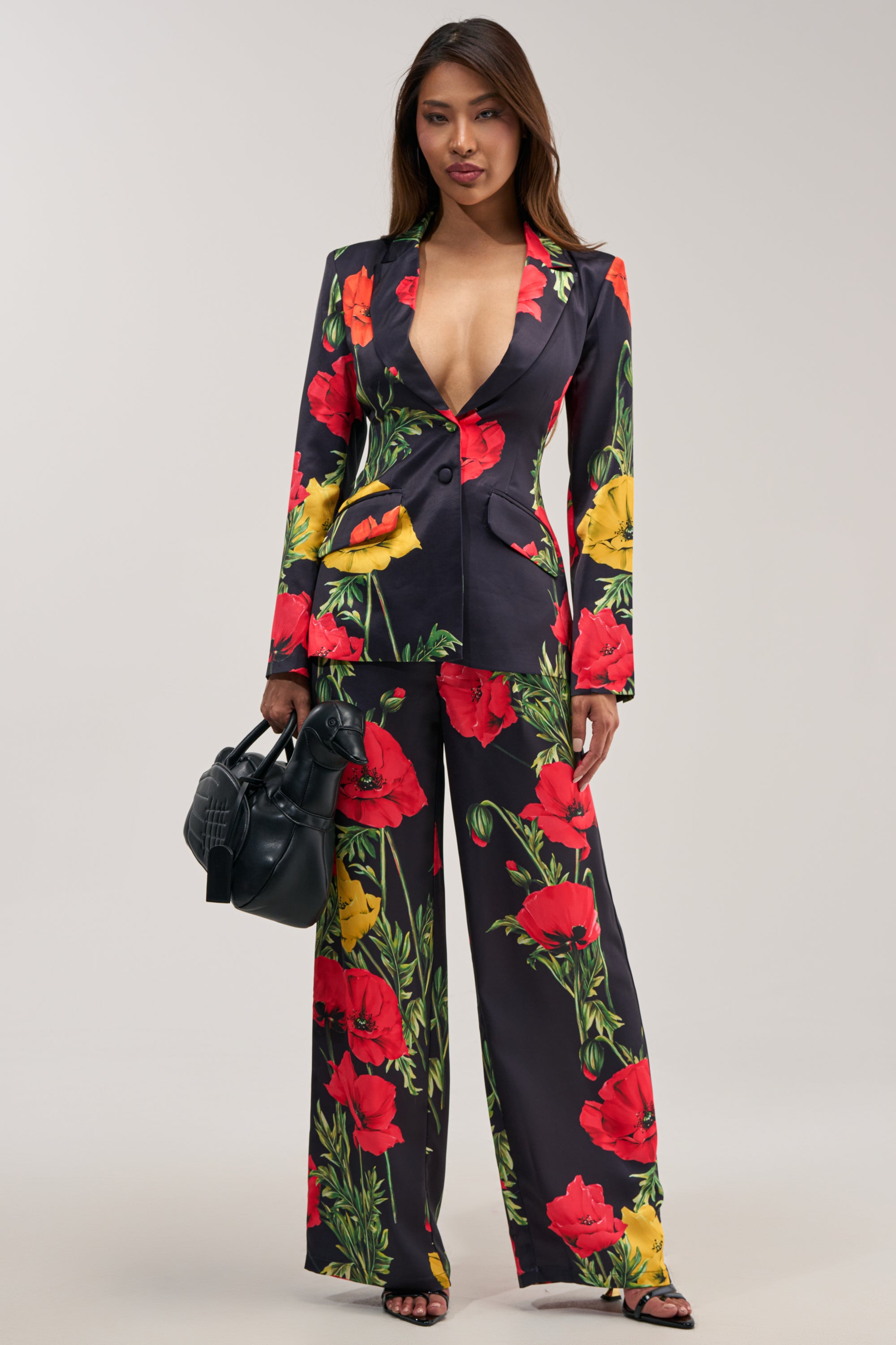 A woman stands confidently in a striking WILDFLOWER SATIN TROUSER set, featuring wide-leg floral print satin trousers adorned with vibrant red and yellow flowers against a dark background. She pairs the bold suit—with its deep neckline—for formal occasions that demand both elegance and statement style, accessorizing with a black handbag and coordinating black sandals.
