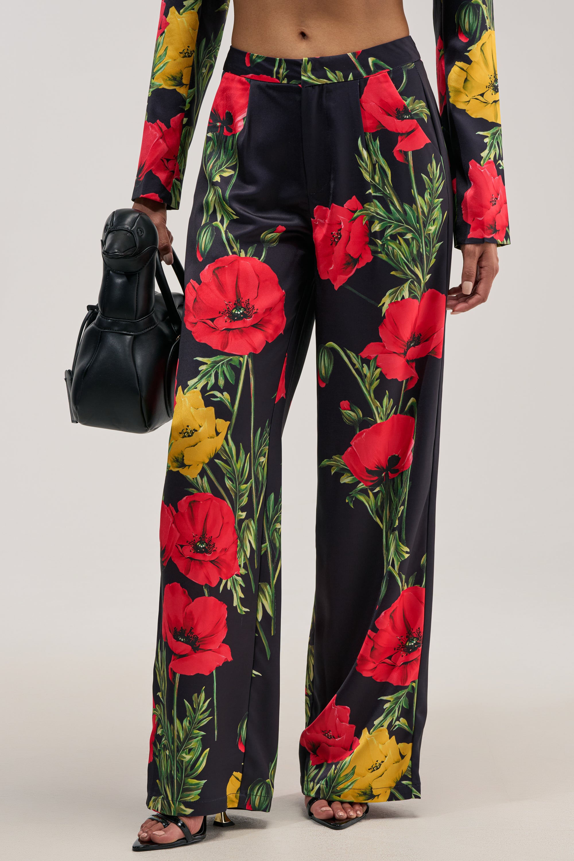 Wearing the WILDFLOWER SATIN TROUSER—black satin trousers adorned with bold red and yellow floral prints and featuring a chic wide-leg silhouette—paired with a black handbag and open-toe heels, this look stands out beautifully against a neutral background, making it perfect for formal occasions.