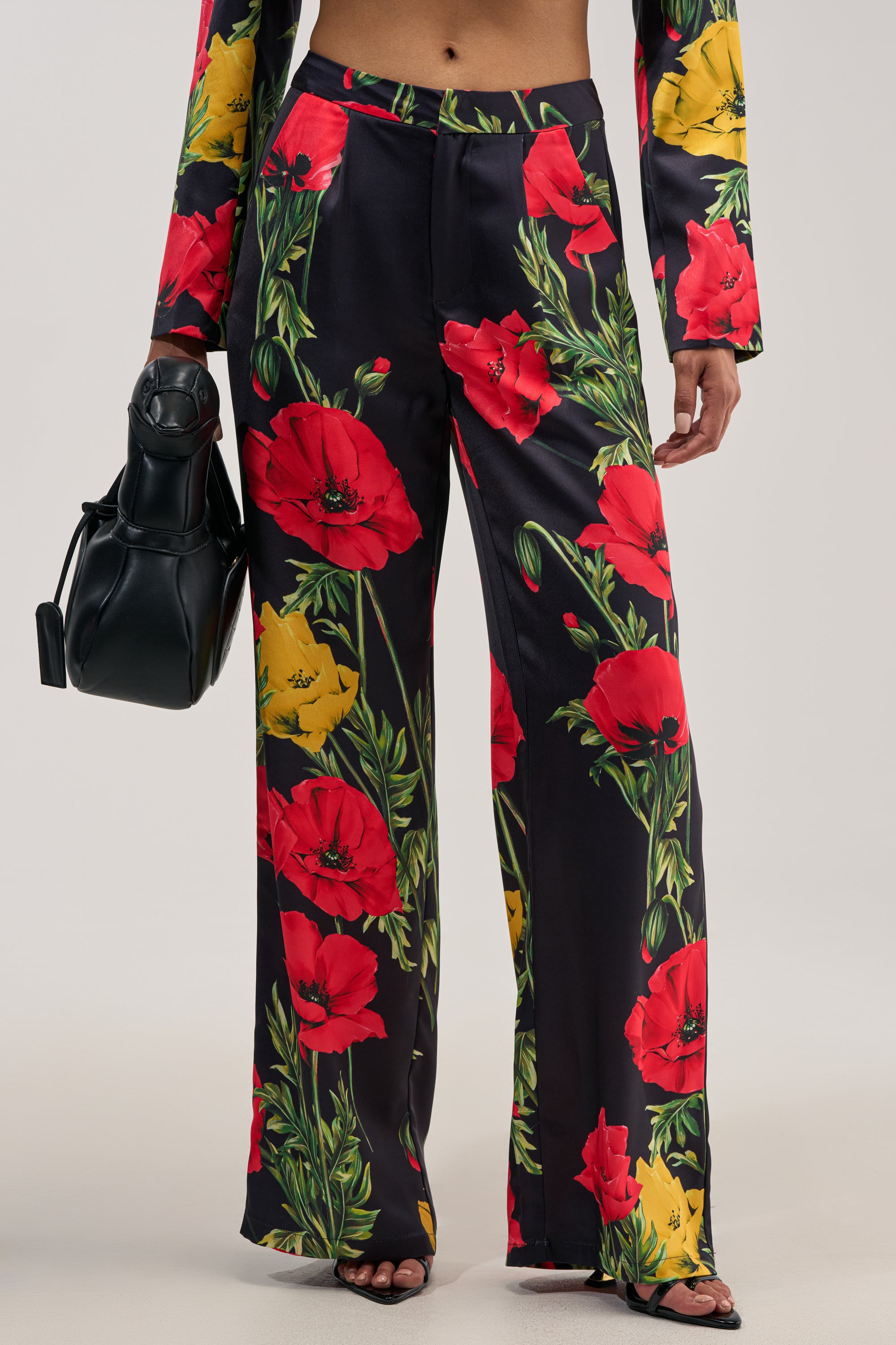 A person wearing the WILDFLOWER SATIN TROUSER—high-waisted satin trousers featuring a vibrant red and yellow floral print and a wide-leg silhouette—paired with a black handbag. The look, showcasing their midriff and open-toed shoes, is ideal for both casual outings and formal occasions.