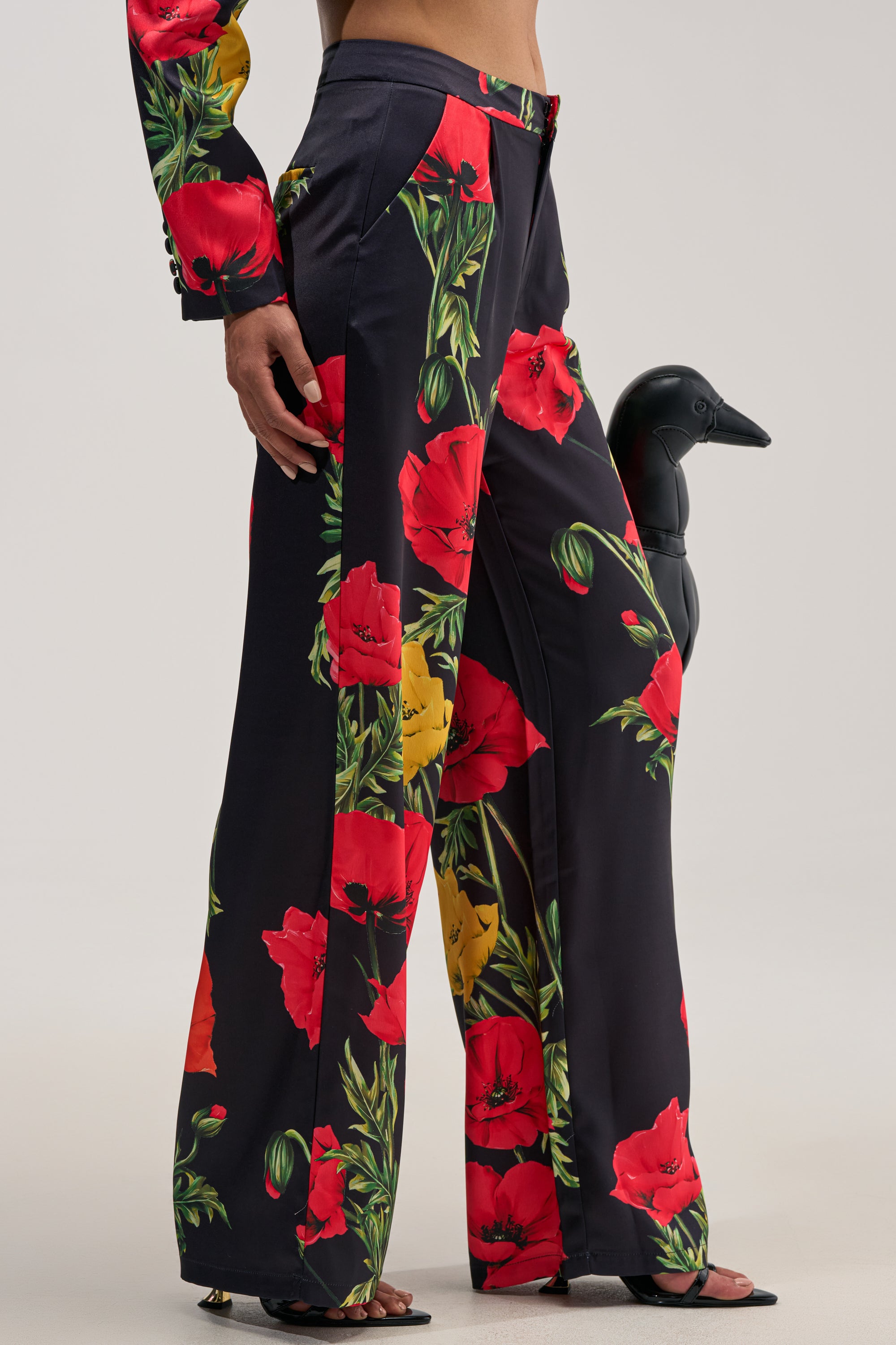 WILDFLOWER SATIN TROUSER in black satin with a wide-leg silhouette, featuring bright red and yellow floral prints; styled with a matching jacket sleeve visible and accessorized with a black bird sculpture against a plain, light-colored background.