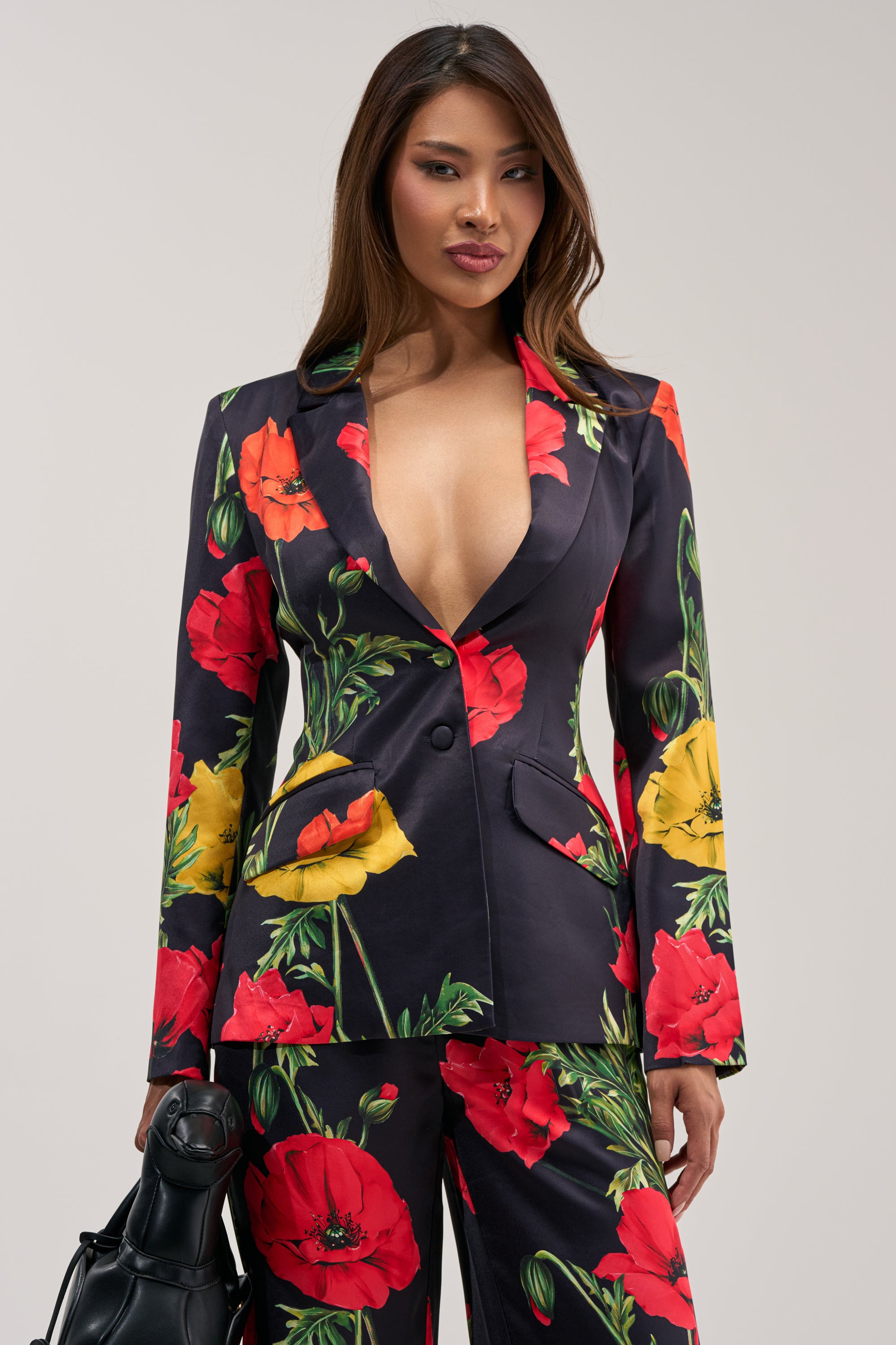 Wearing the WILDFLOWER SATIN BLAZER featuring a deep V-neck and vibrant red, yellow, and green florals, she holds a black handbag against a plain light background—a stunning choice for your next summer date night.