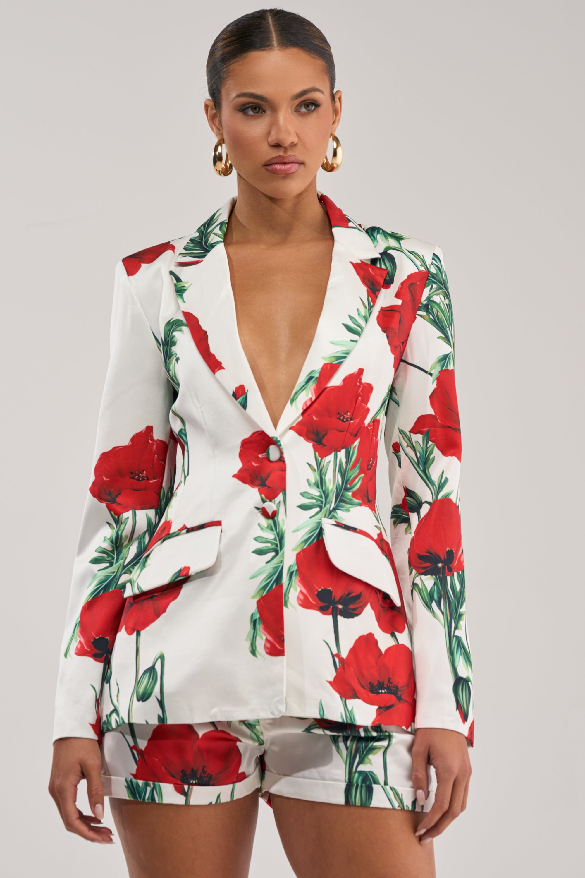 A woman wearing the POPPY SATIN BLAZER with a floral print, matching shorts, gold hoop earrings, and slicked-back hair stands against a plain light gray background.