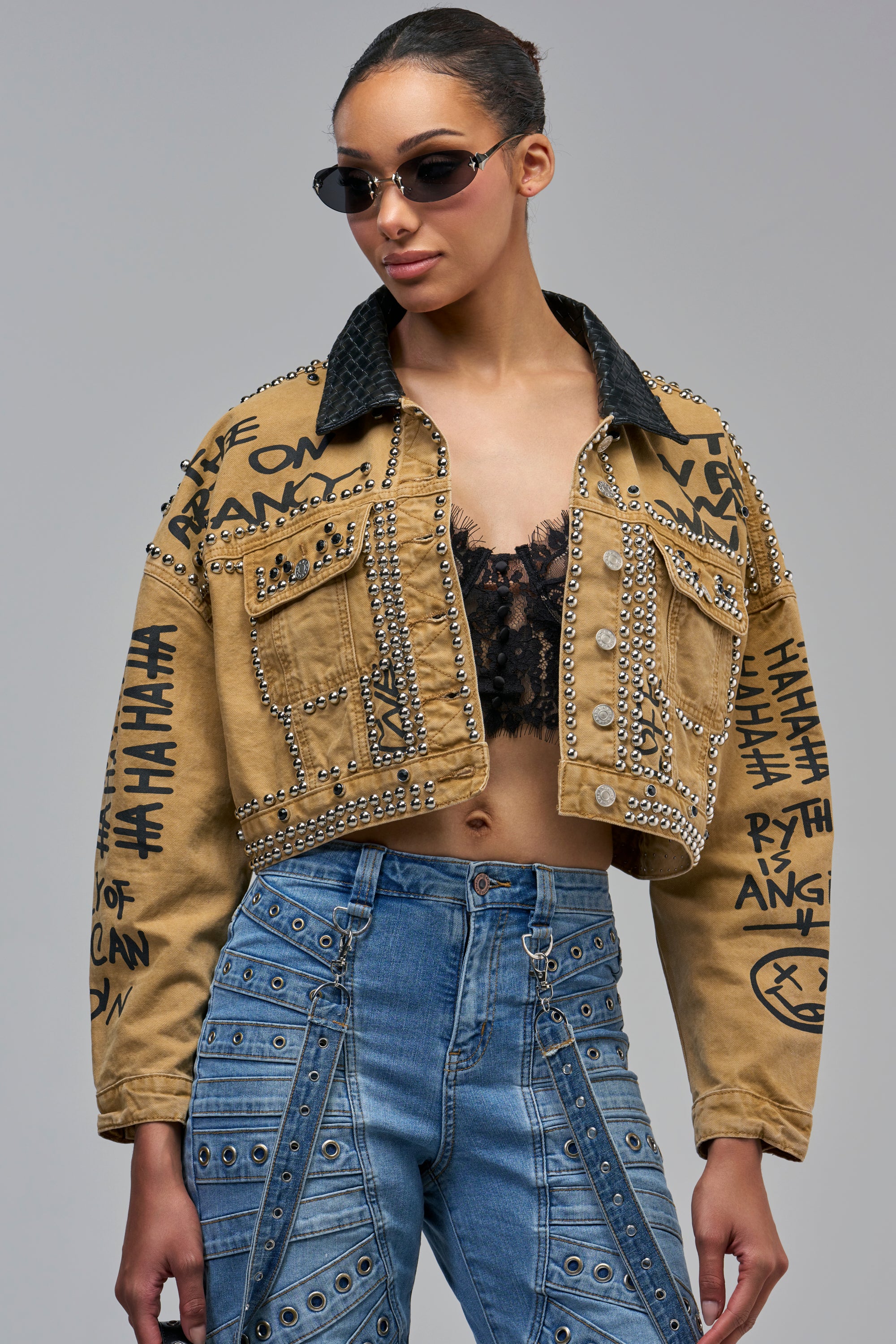 A cropped khaki BANDIT STUDDED UTILITY JACKET featuring black graffiti text, silver studs, and a black collar, worn over a black lace bralette.