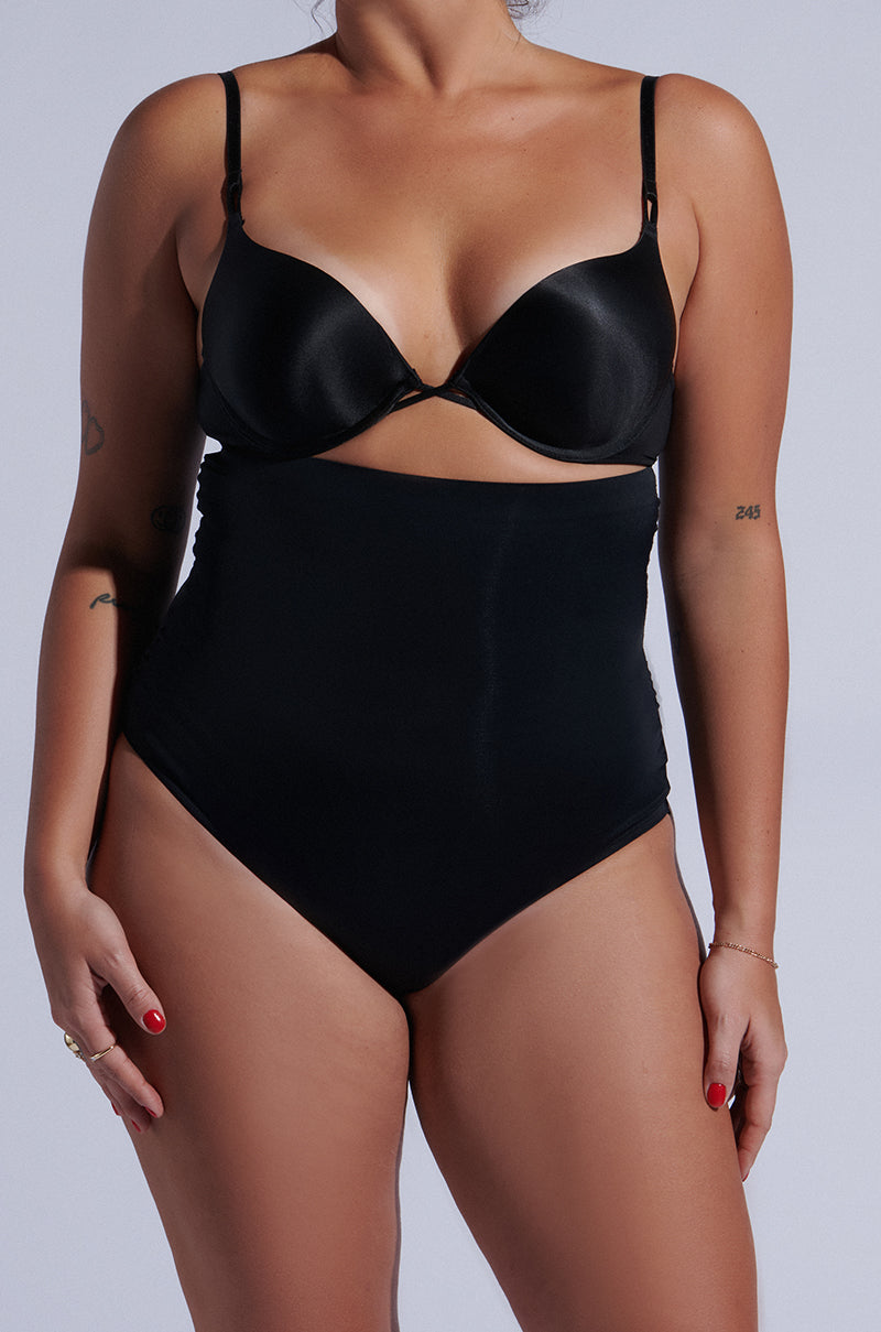 Wearing the Titsy Shaper Thong in Black and high-waisted shapewear with seamless hems, the person stands confidently against a neutral background. Their tattooed arms and vibrant red fingernails are complemented by a bracelet on one wrist.
