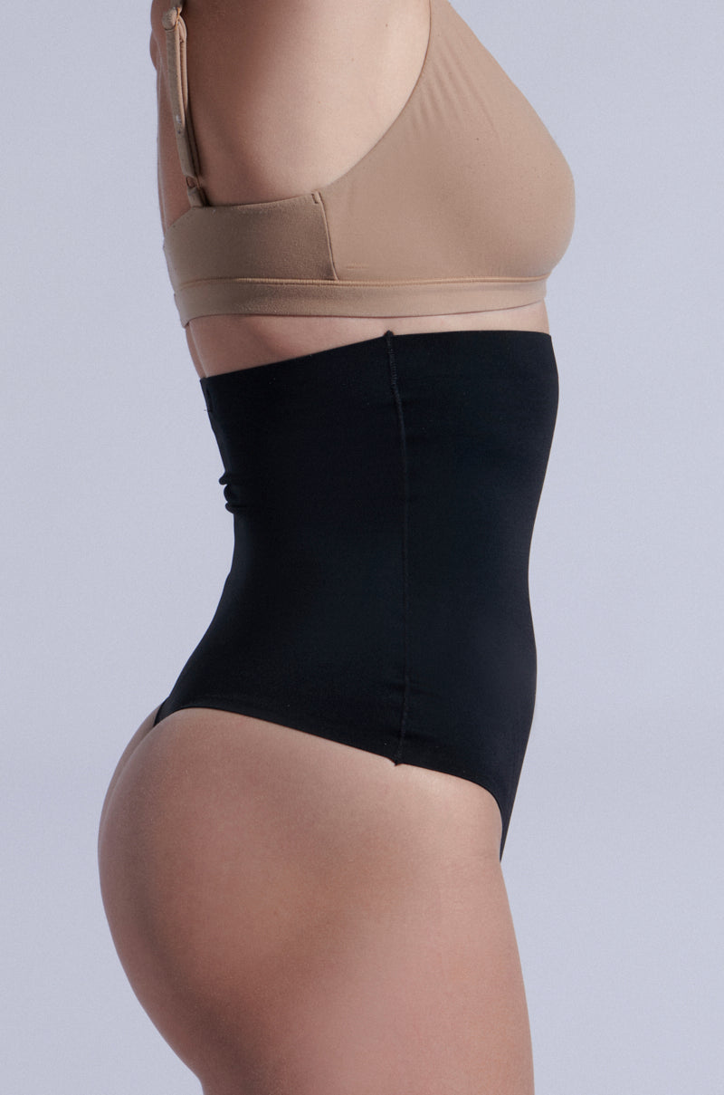 A person models the TITSY Shaper Thong in black, highlighting its seamless design and high-rise fit from a side profile against a light gray background, paired with a nude-colored bra.