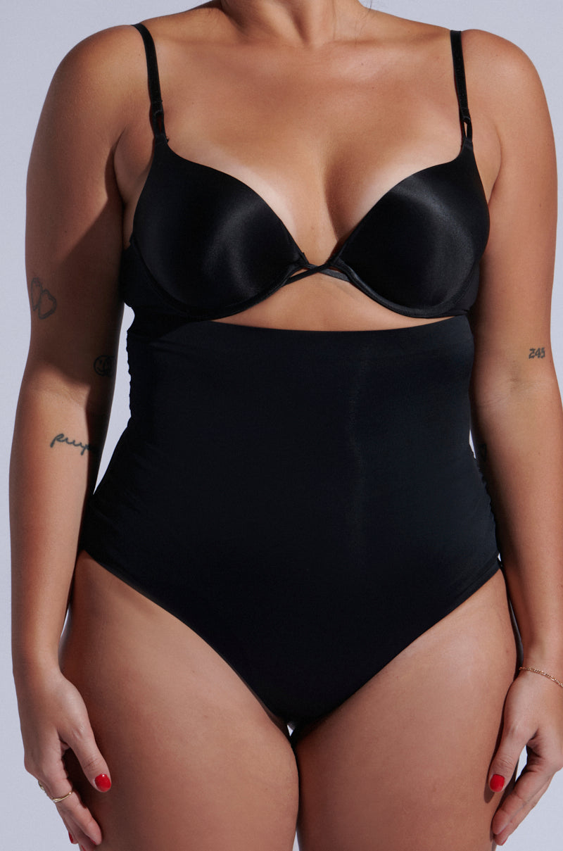 A person is seen from the front wearing the TITSY SHAPER THONG IN BLACK, featuring a high-rise fit with matching shapewear panties and bra. The look is completed by seamless hems, with small tattoos visible on their arms.