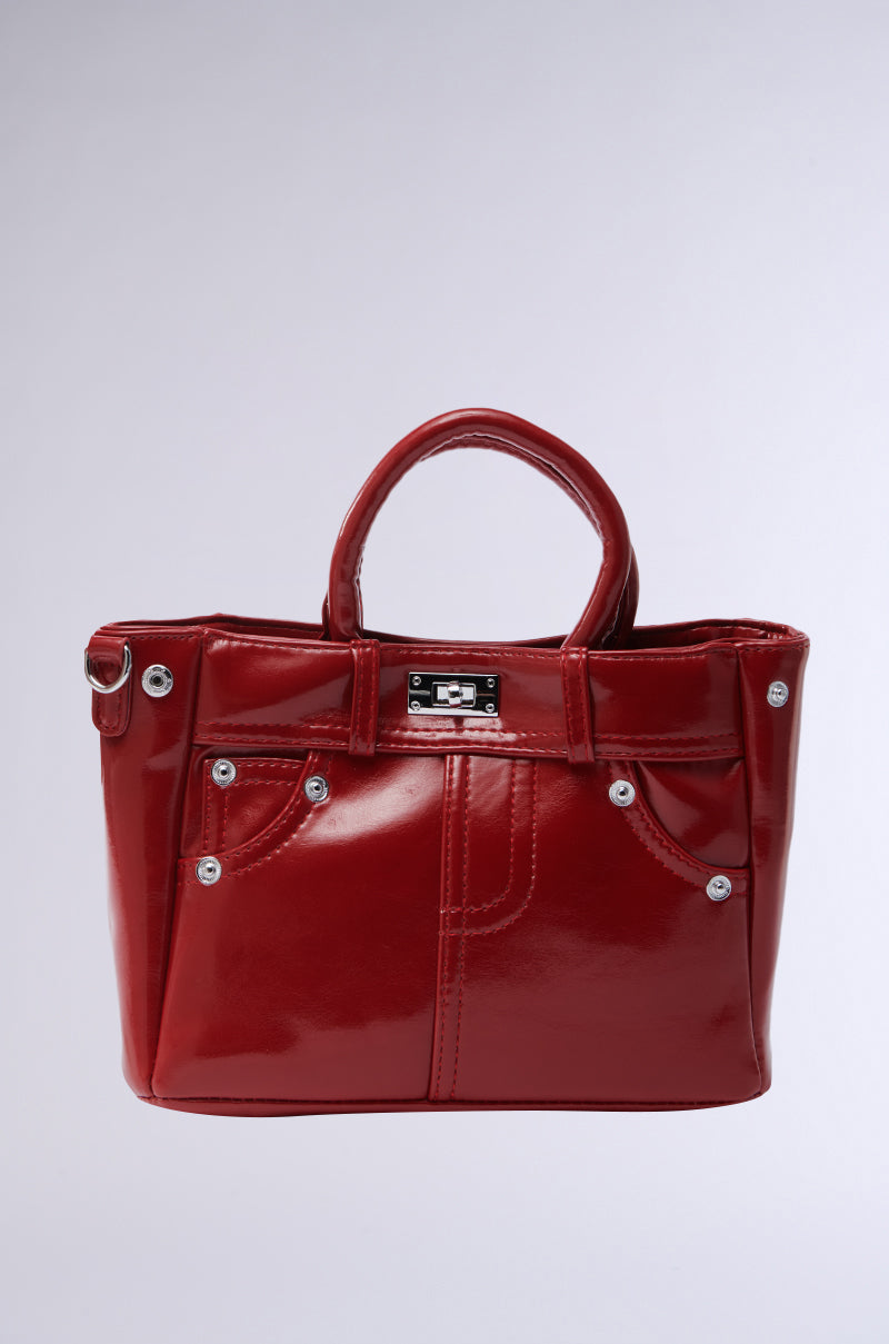 The glossy red patent leather "TOP TIER PANTS PU PURSE" features two handles, decorative stitching, silver rivets, and a twist-lock top closure with a small metal plate. Its vibrant color and intricate details stand out against a plain background.