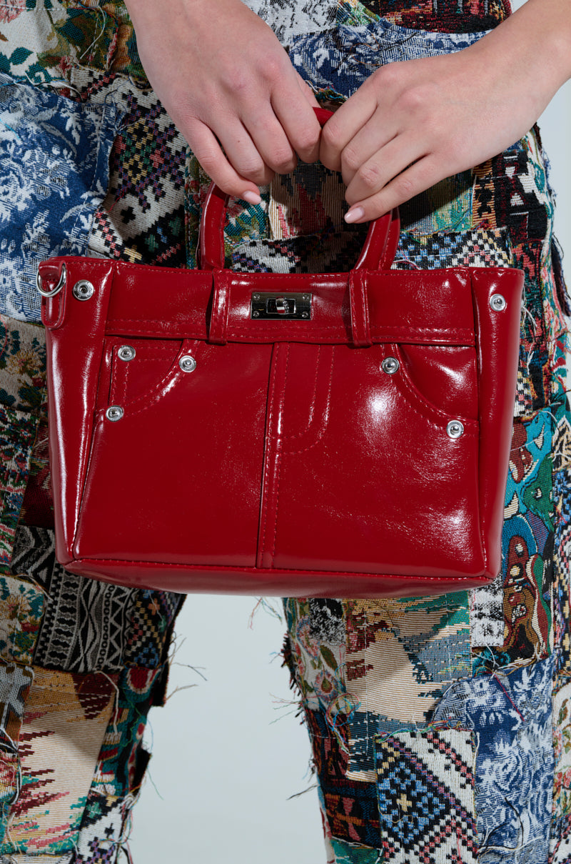 A person carries the TOP TIER PANTS PU PURSE, showcasing shiny red patent leather with metal accents and an adjustable strap, while donning vibrant patchwork pants that boast floral and geometric patterns.