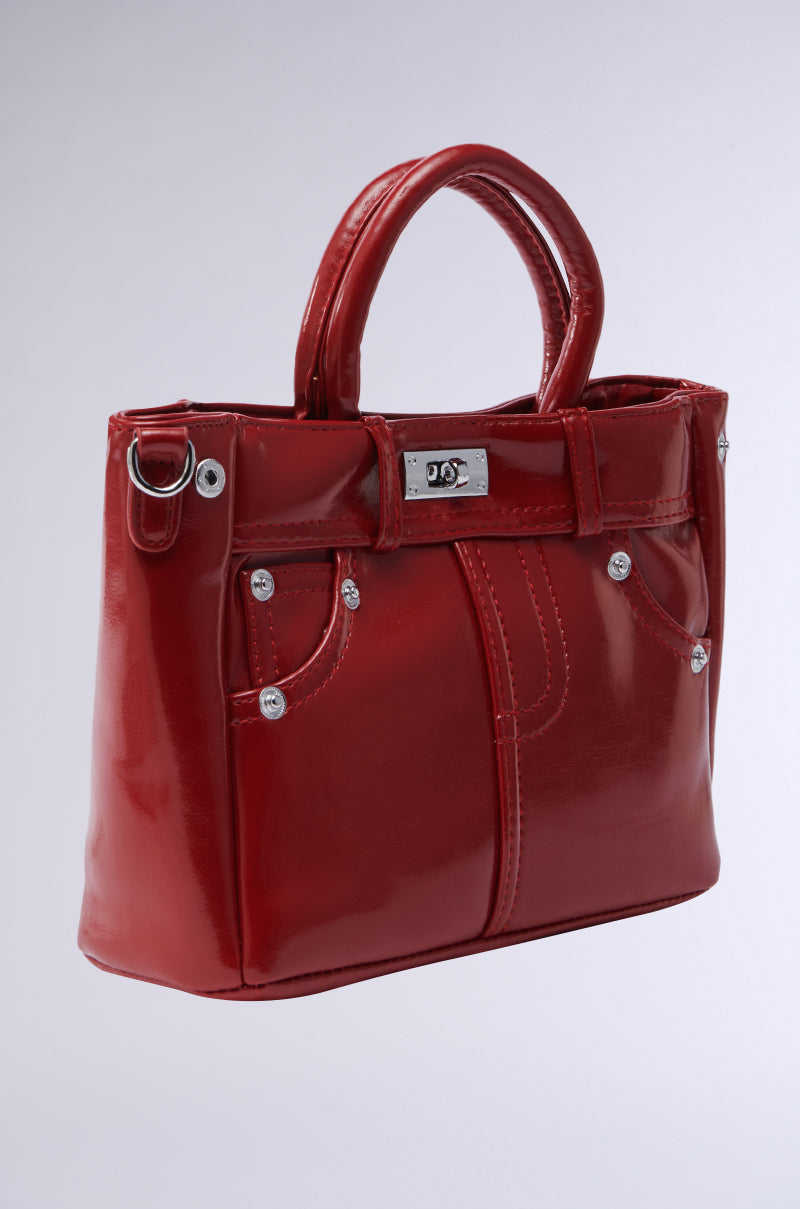 The TOP TIER PANTS PU PURSE is a shiny red patent leather bag with a glossy finish, featuring two handles, decorative stitching, a twist-lock closure, an engraved metal plate on top, button-like embellishments, and two subtle front pockets.