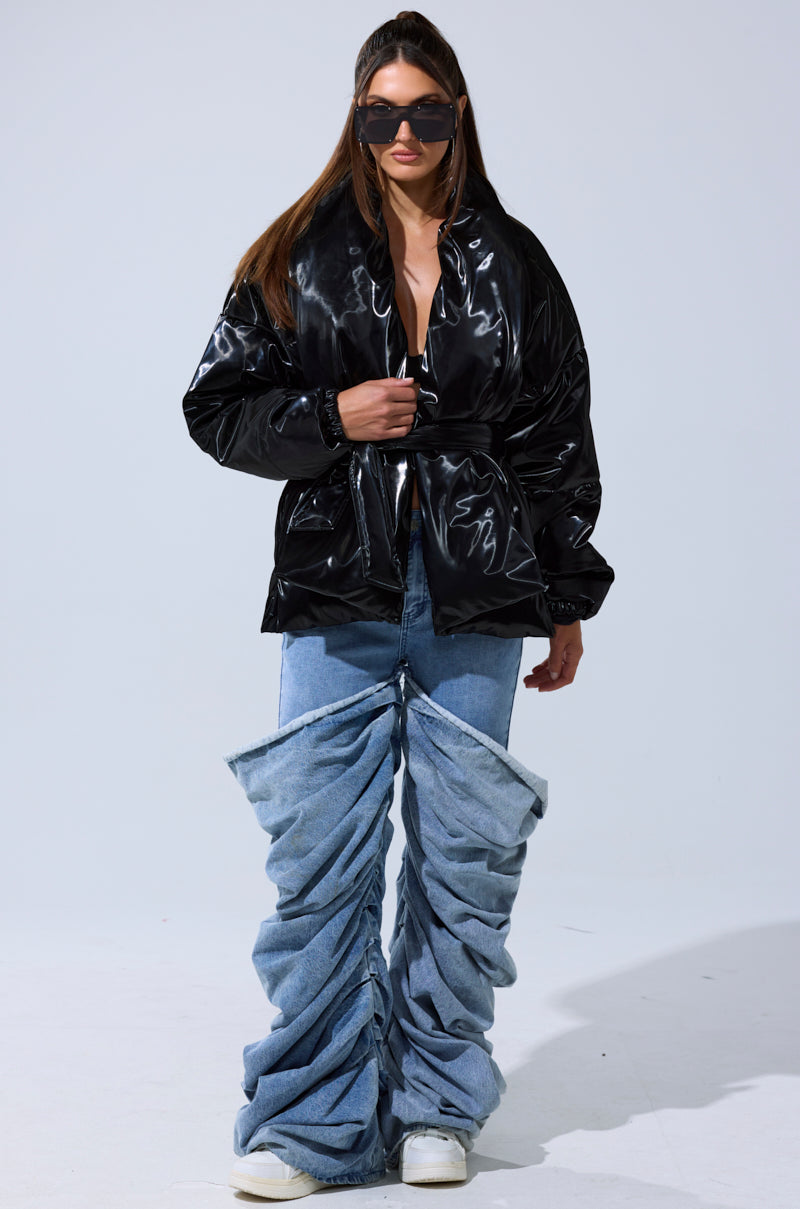 A woman in the LIQUID METAL WRAP PUFFER pairs it with oversized ruched blue jeans, white sneakers, and large black sunglasses while standing against a plain light background.