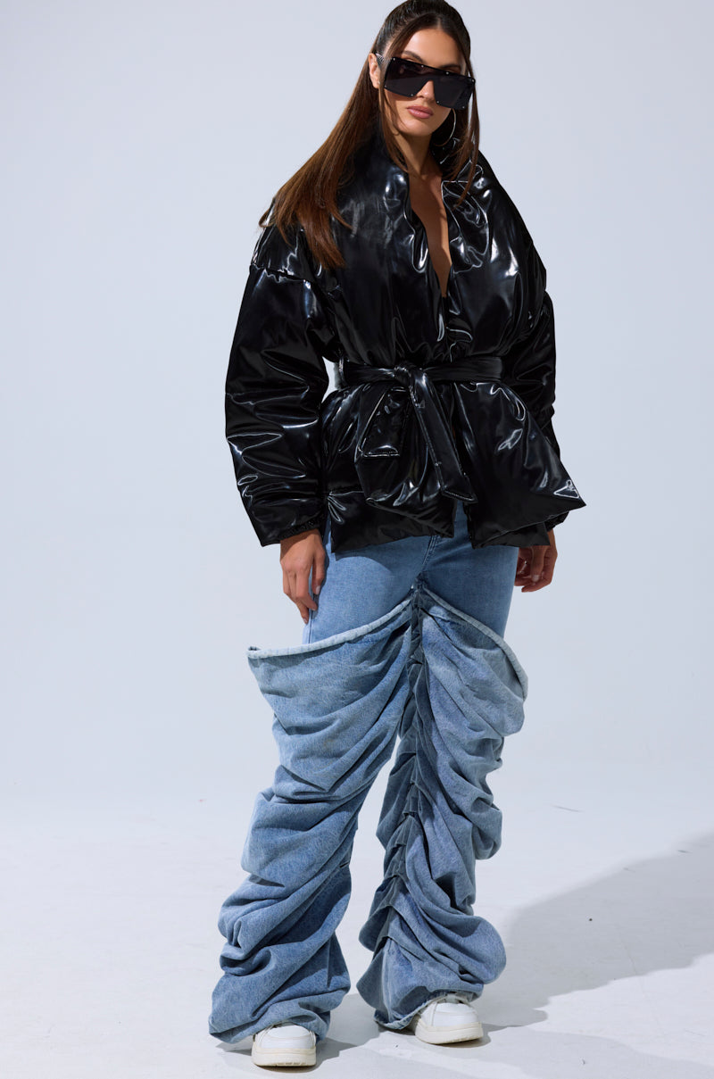A woman poses in the LIQUID METAL WRAP PUFFER, oversized ruched denim jeans, white sneakers, and large black sunglasses against a plain light background.
