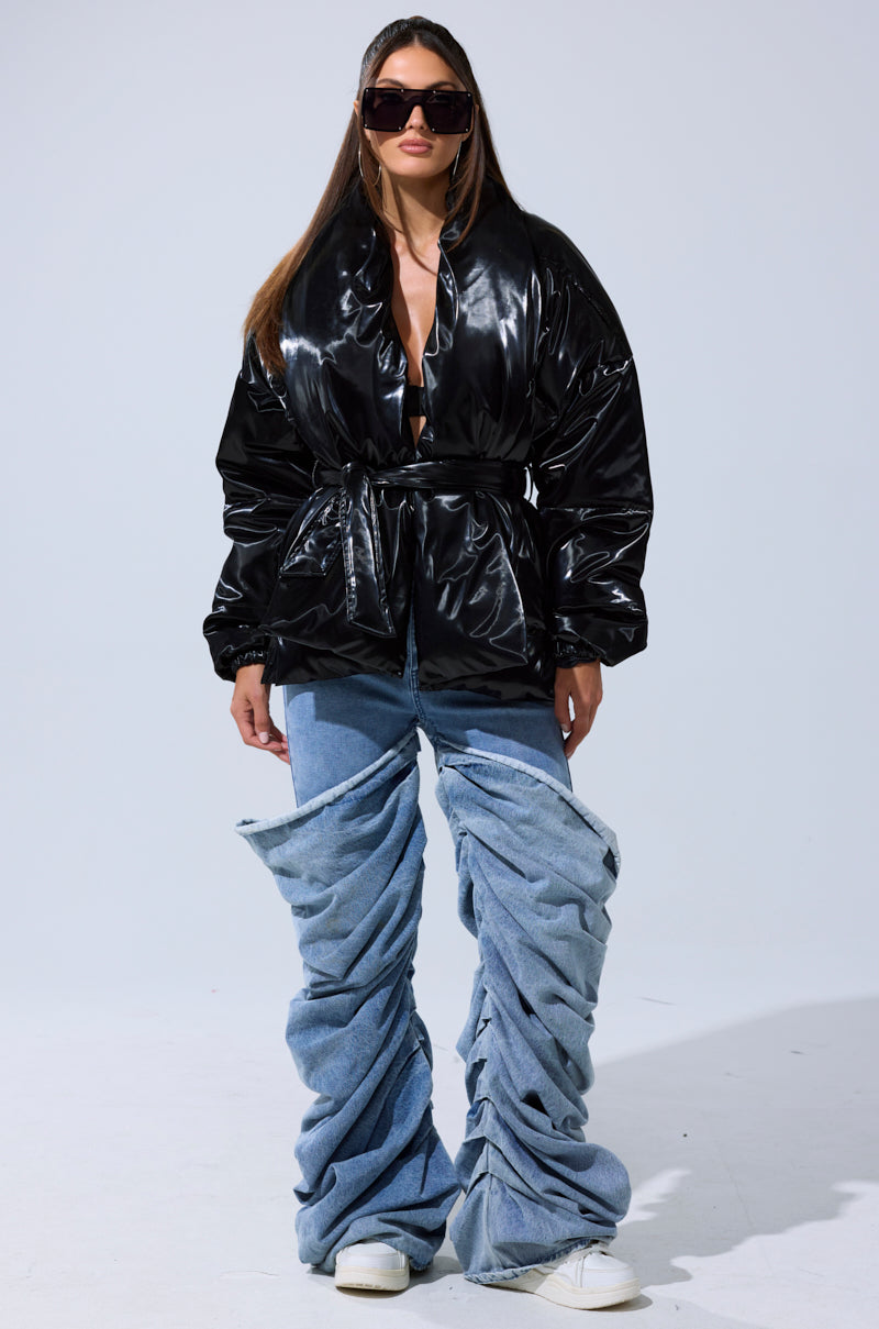 A woman in oversized sunglasses, the LIQUID METAL WRAP PUFFER with a belted waist, and baggy ruched blue jeans over white sneakers stands against a plain backdrop—effortlessly exemplifying chic outerwear style.