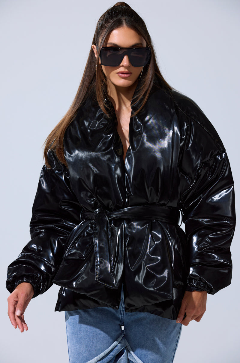 A woman poses confidently against a plain light background, wearing the LIQUID METAL WRAP PUFFER, styled with a belt, light blue jeans, and large black sunglasses.
