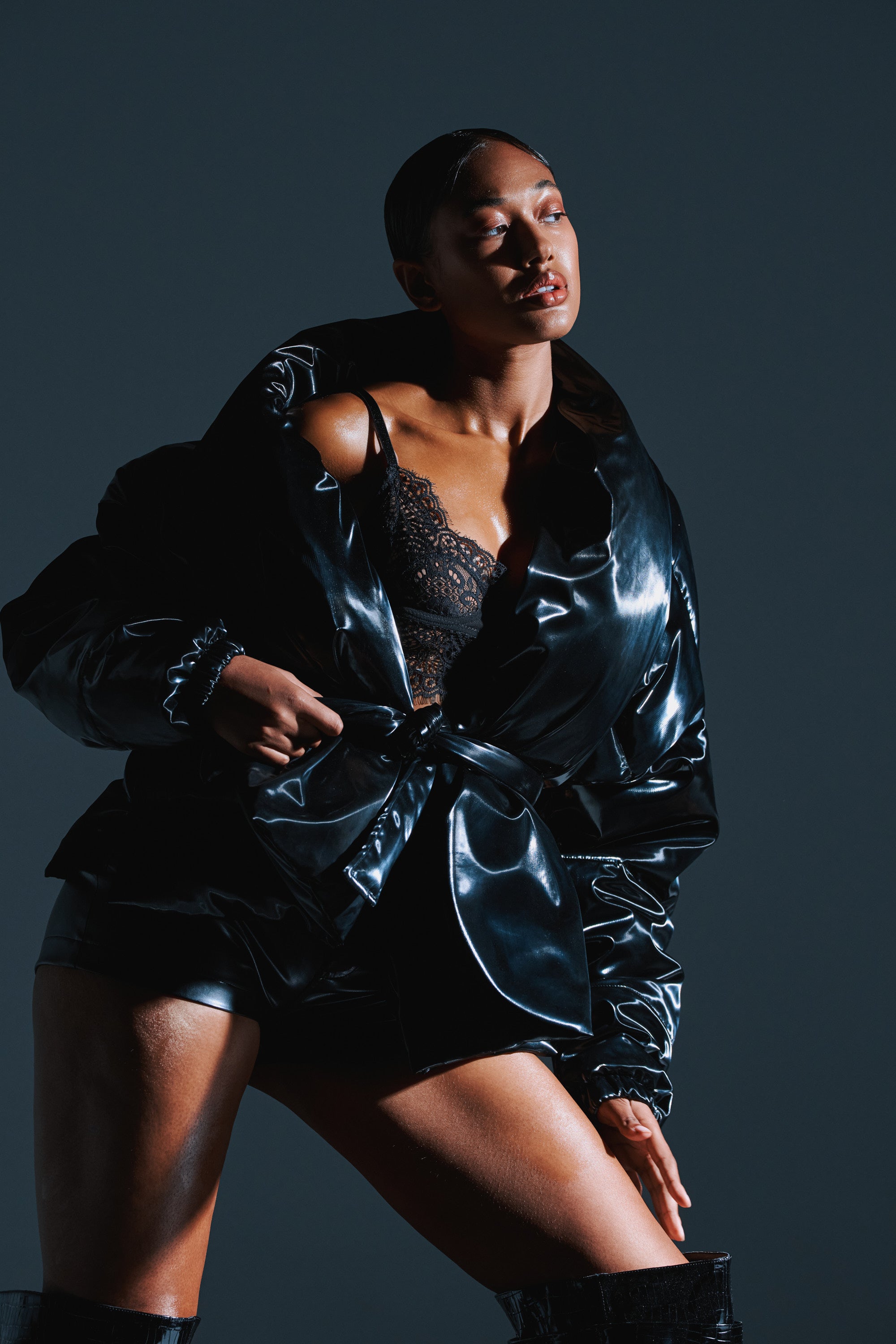 A woman poses confidently in the LIQUID METAL WRAP PUFFER—a glossy black jacket—styled over a lace top and shorts against a dark background. Her slicked-back hair and upward gaze are highlighted by dramatic lighting.