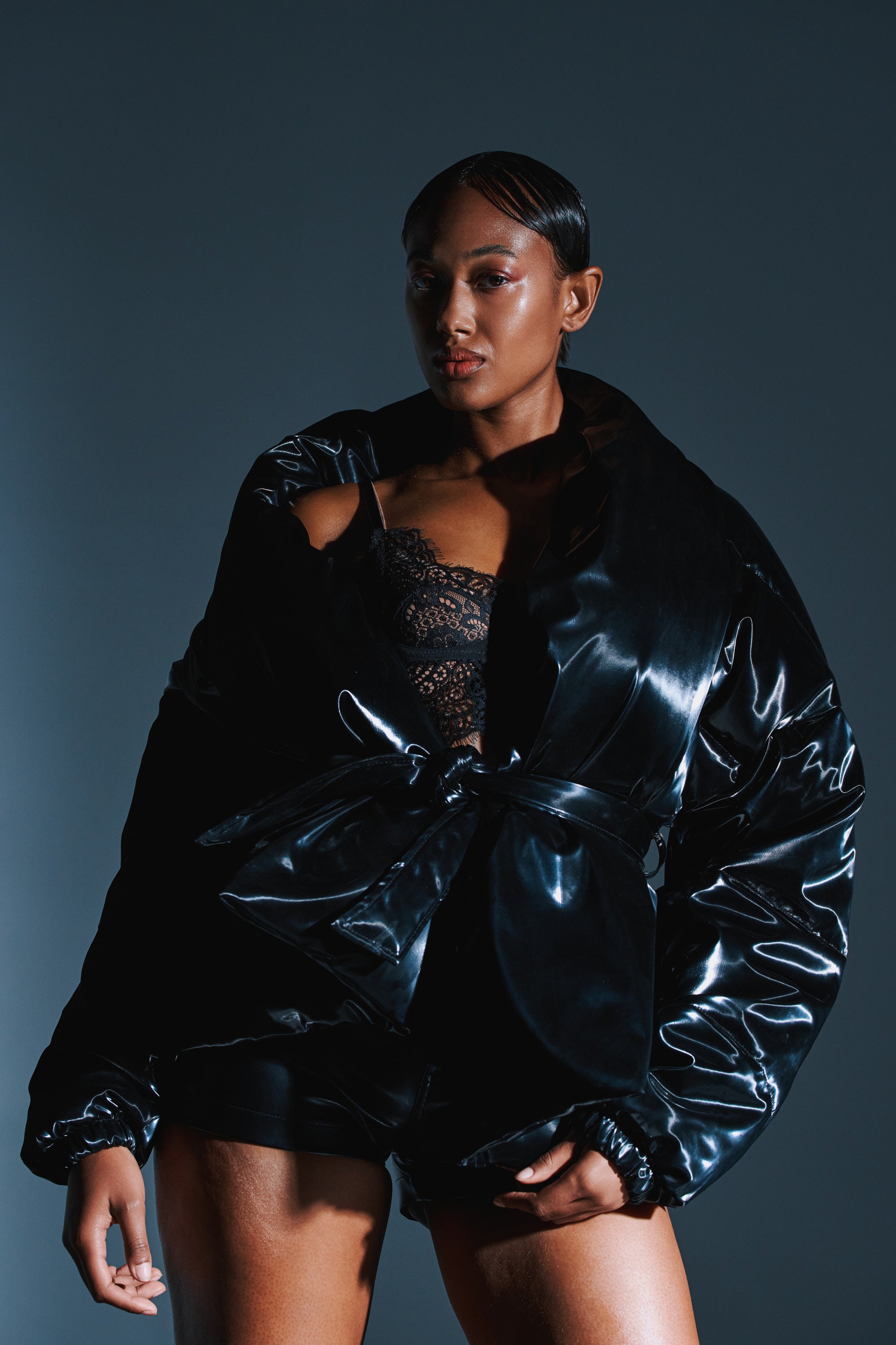 A woman poses confidently in the LIQUID METAL WRAP PUFFER, an oversized shiny black jacket belted at the waist over a black lace top and shorts, with slicked-back hair against a dark background.