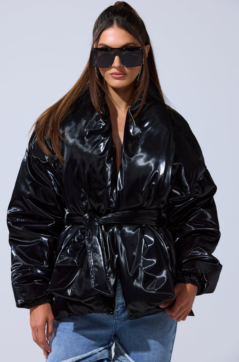 A woman stands against a plain light background wearing the LIQUID METAL WRAP PUFFER with a belt, paired with light blue ripped jeans and oversized black sunglasses.
