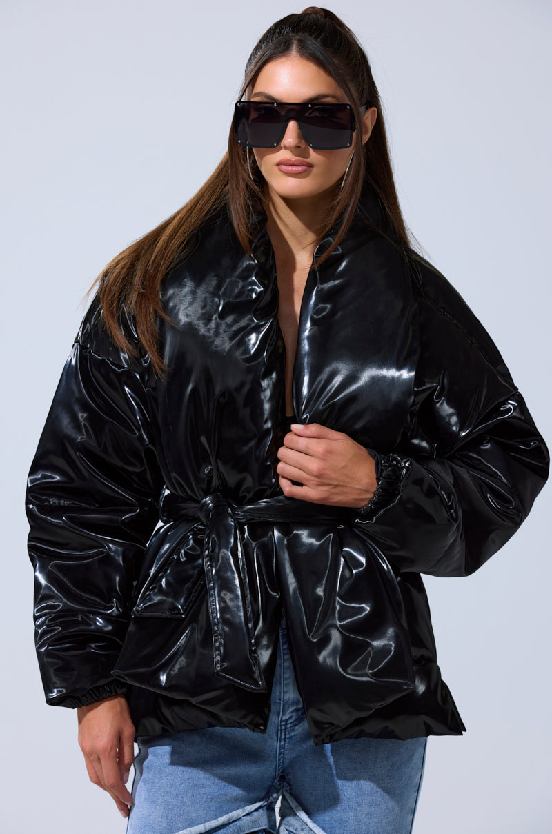 A woman with long, straight brown hair wears the LIQUID METAL WRAP PUFFER with a waist belt, paired with oversized black sunglasses and ripped blue jeans, posing confidently.