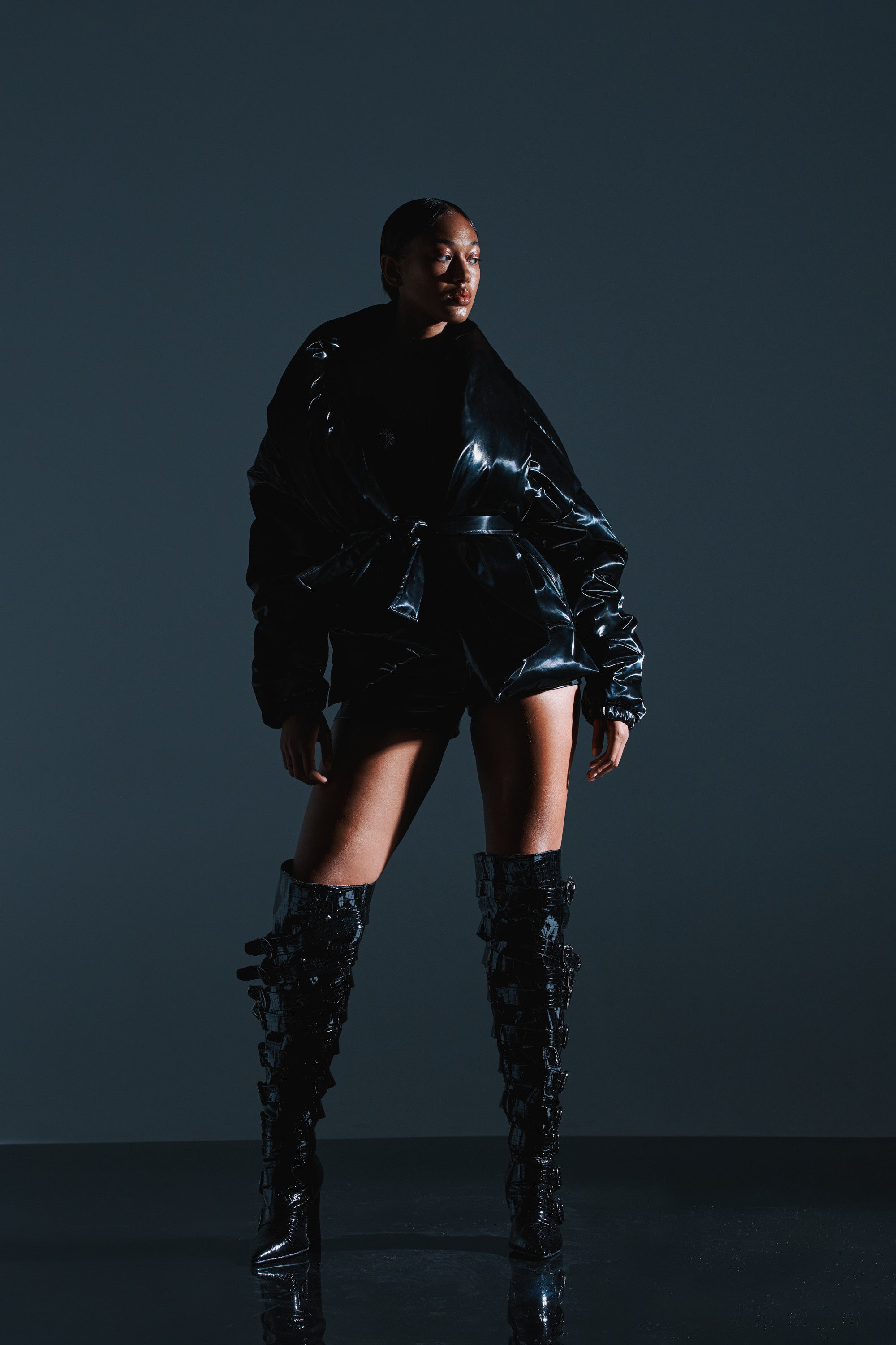 A person stands confidently in a dark studio, wearing the LIQUID METAL WRAP PUFFER, matching shorts, and knee-high black patent leather boots with buckles. The lighting highlights the jacket's glossy winter texture.