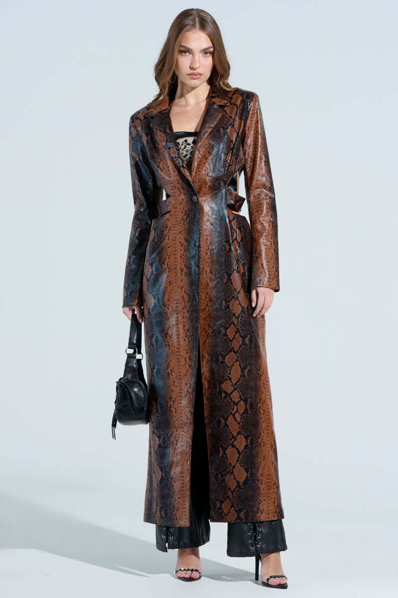 A woman confidently poses in the MORE LIFE CUTOUT FAUX LEATHER TRENCH over black pants, holding a small black handbag. Her loose hair complements the look against a plain, light background.