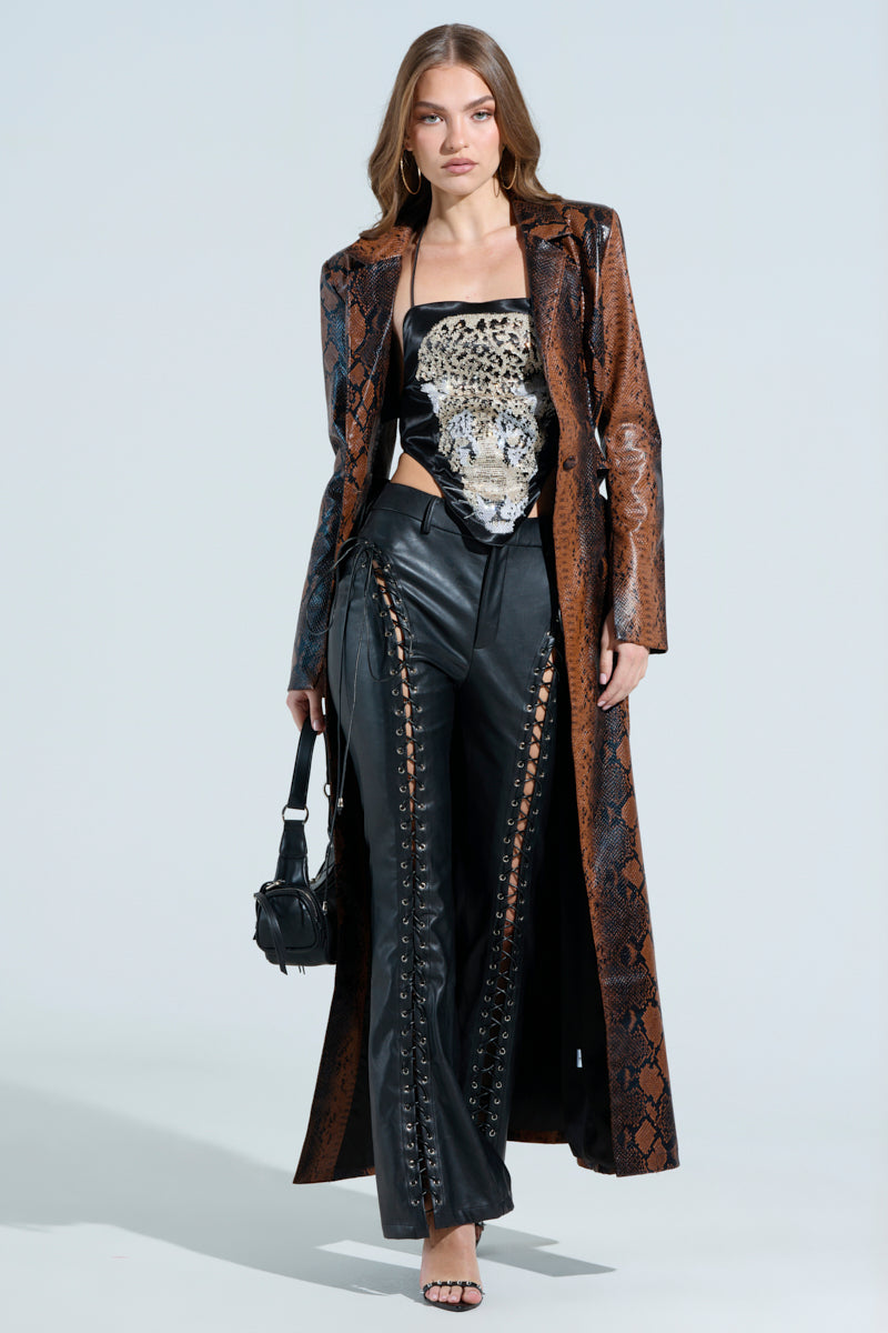 A woman poses confidently in the MORE LIFE CUTOUT FAUX LEATHER TRENCH over a leopard print top. She wears black leather lace-up pants, holds a small handbag, and has long hair styled loosely, standing against a plain background.